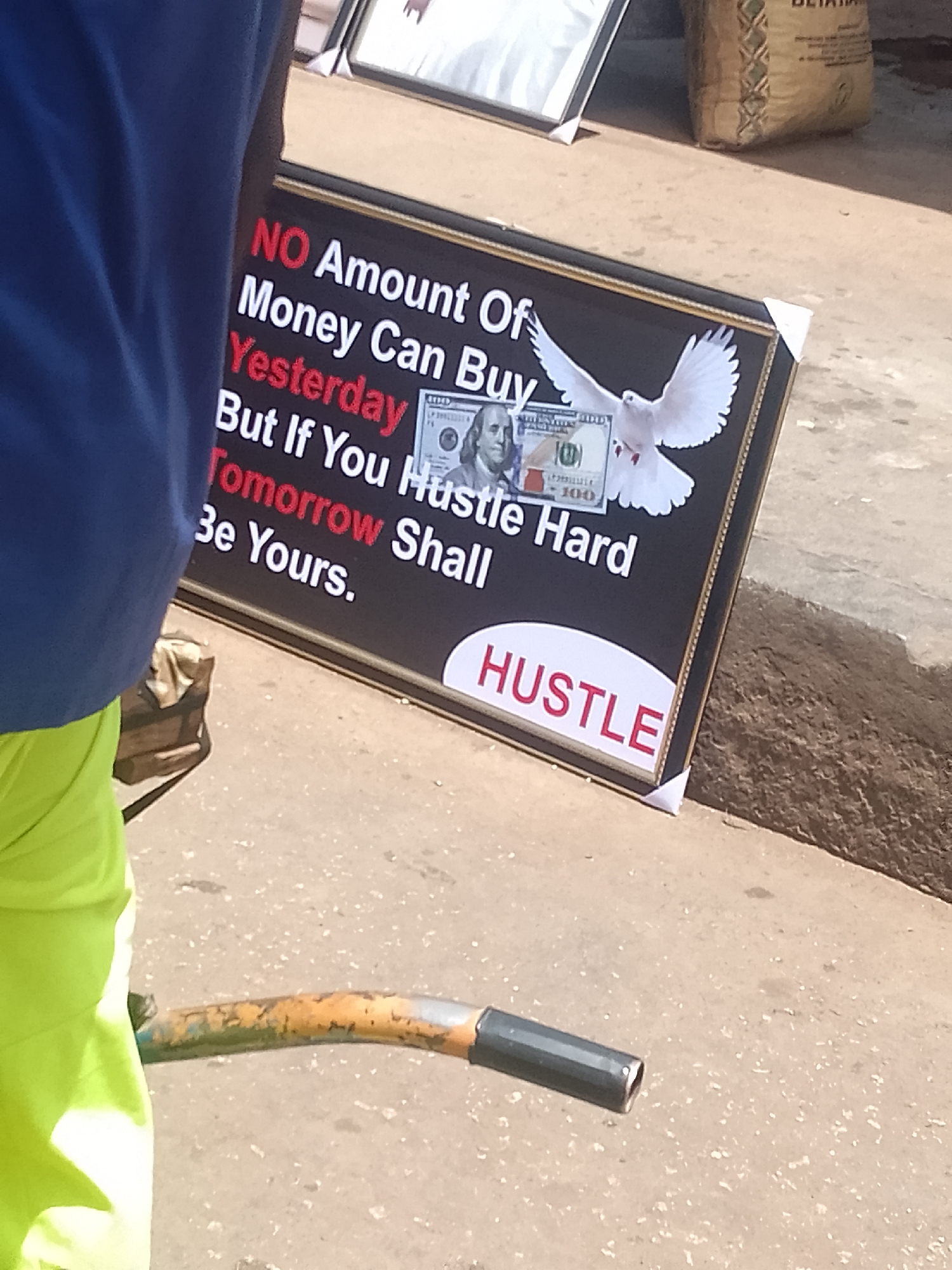 The image shows a framed poster with a motivational message, likely located in Bukuru, Nigeria. The poster reads: "NO Amount Of Money Can Buy Yesterday But If You Hustle Hard Tomorrow Shall Be Yours." The word "HUSTLE" is prominently displayed in red letters at the bottom. The poster also features an image of a US $100 bill with a white dove. Parts of two people are visible. The person on the left wears bright green shorts and a blue shirt. In the upper part of the picture is part of a bag of flour and another framed picture.