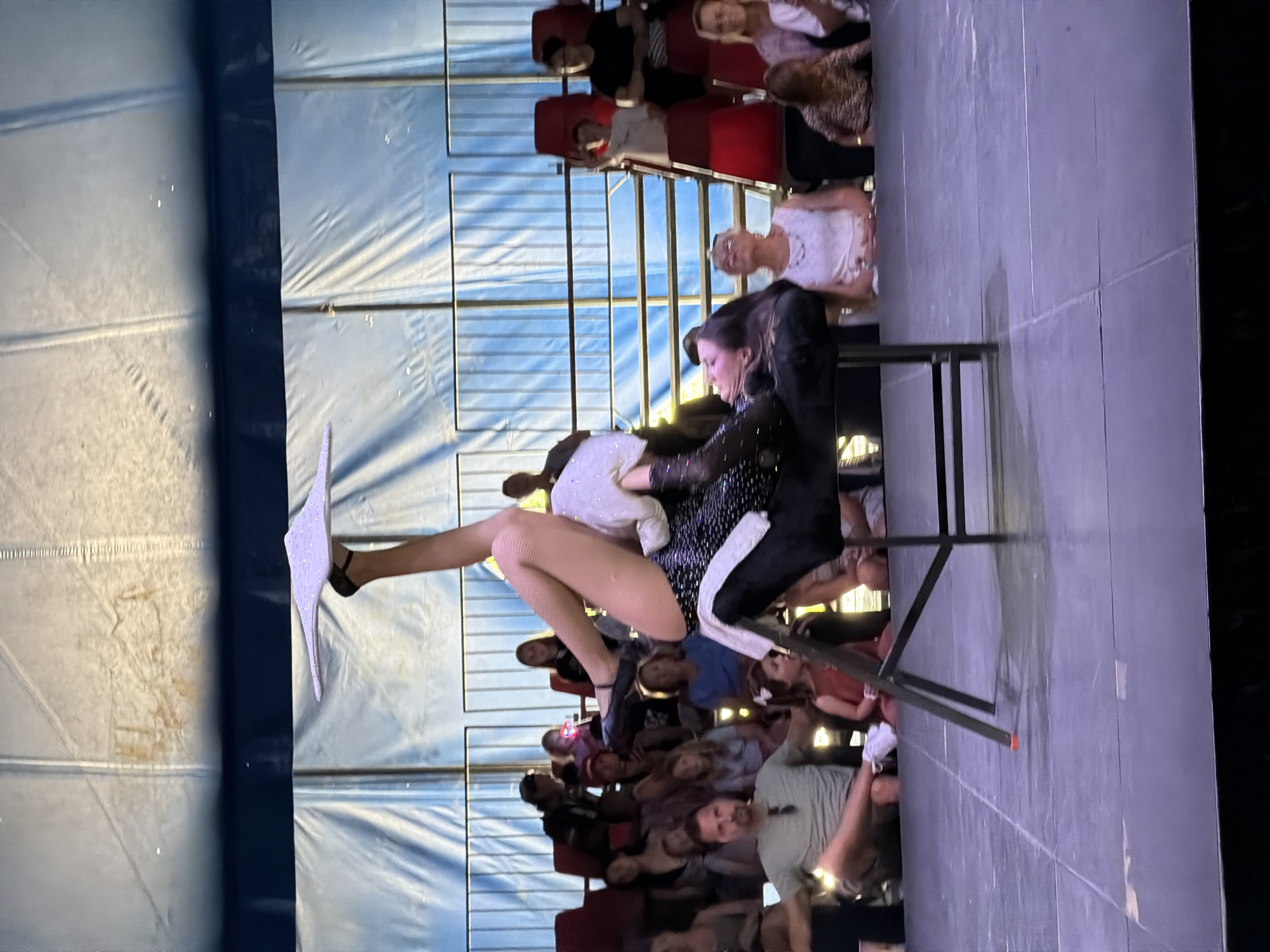 An aerial performer balances an object on her foot while lying on a chair on a stage. The performer, dressed in a sparkly black outfit and fishnet stockings, is positioned on her back with her legs extended upwards. The object on her foot appears to be a flat, light-colored disc with a speckled surface. The stage is a dark, flat surface with visible seams. Behind the stage, an audience is seated in tiered rows of red chairs. The backdrop consists of blue fabric draped across a structure. The lighting suggests an outdoor evening event. The location is the Fort Erie Race Track in Fort Erie, Canada.