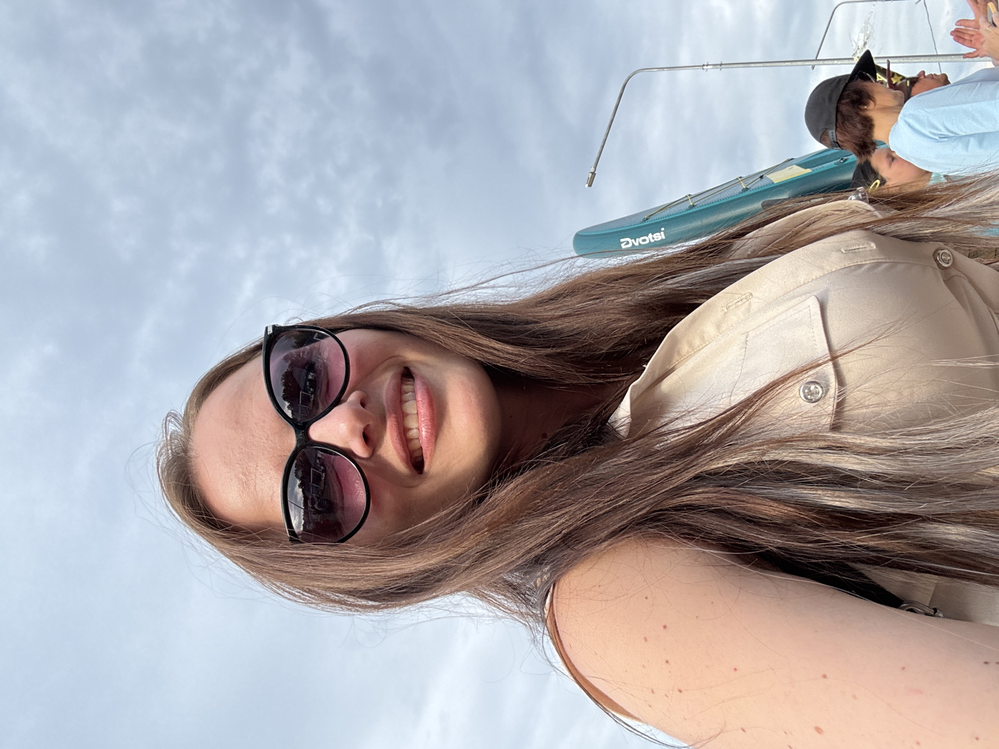 A woman with long brown hair and sunglasses smiles at the camera outdoors. She is wearing a light-colored sleeveless shirt. In the background, a turquoise object with "Dvotsi" written on it is visible, along with a street lamp and other people. The sky is cloudy. The image appears to be taken in Chaikovsky, Russia, based on the presence of the "Dvotsi" brand, which is associated with a regional bus company in that area.