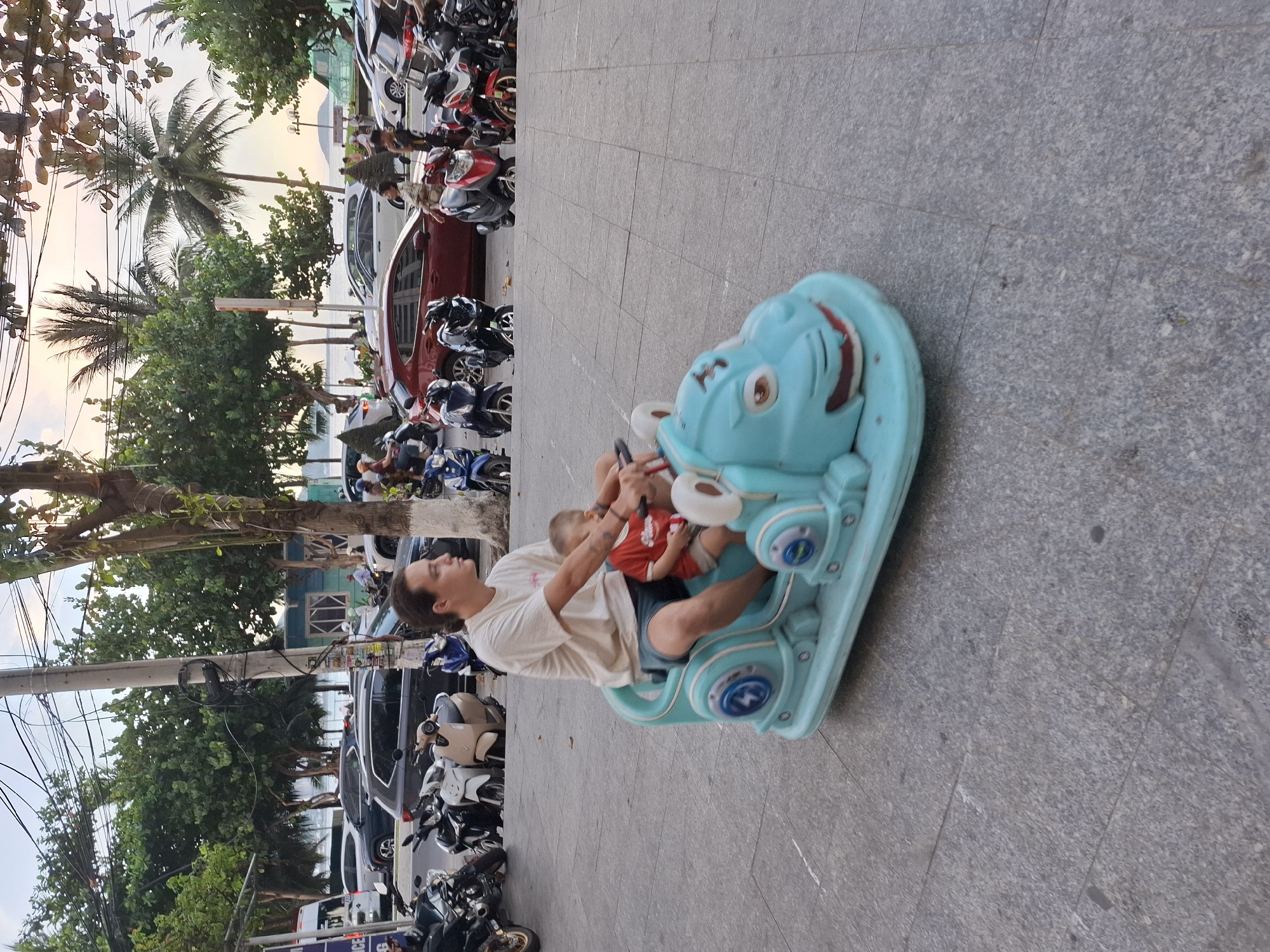 A man and a young child are sitting on a blue rocking toy on a paved walkway. The man is wearing a white t-shirt and blue shorts, and the child is wearing a red shirt. They are in the Bac Nha Trang Ward, Vietnam. In the background, there are many parked motorcycles and scooters, as well as trees and a body of water that appears to be the ocean. The time of day appears to be late afternoon or early evening, as the sky is a soft orange and pink. The overall mood is relaxed and peaceful, with the man and child enjoying their time together.