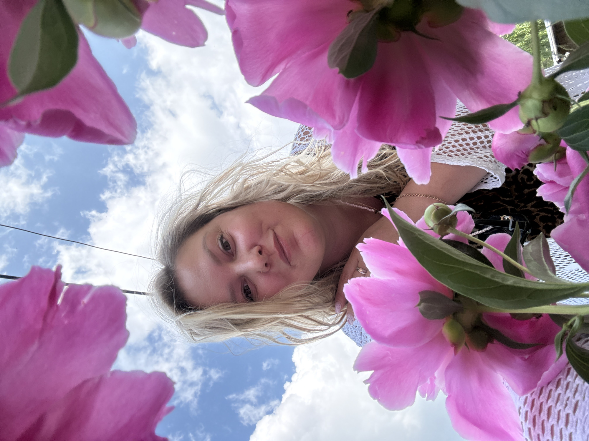 This is a close-up, low-angle outdoor shot of a woman framed by large, pink peony flowers. The woman, with blonde wavy hair, is looking towards the camera with a slight smile. She is wearing a white, crocheted garment over a dark, patterned top. A thin necklace is visible. Her hands are visible, one resting near a peony. The background consists of a cloudy blue sky. The composition suggests the photo was taken on a sunny day, possibly in a garden or park setting. There is no information to suggest this scene is in Chaikovsky, Russia.