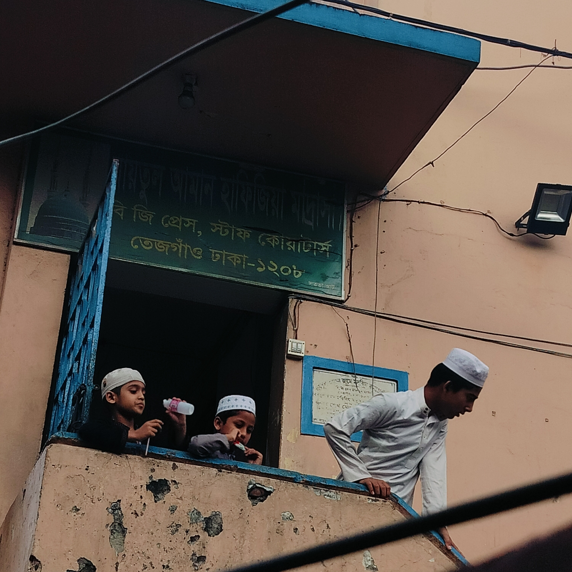 Three young boys are visible in the image, seemingly hanging out in the window of a building. Two of the boys are leaning on the windowsill, wearing what appear to be Muslim skullcaps. One is holding a clear plastic water bottle. A third boy, also wearing a cap and a white shirt, is leaning over the side of a railing next to the window, facing to the right.
A sign in green lettering above the window reads "Baitul Aman Hafizia Madrasa, BJ Press, Staff Quarters, Tejgaon Dhaka-1208", indicating the building is a Madrasa (Islamic school) named Baitul Aman Hafizia, located in the Tejgaon area of Dhaka, Bangladesh. A lamp is mounted on the wall to the right. The building exterior is a peach color with a weathered and peeling texture.