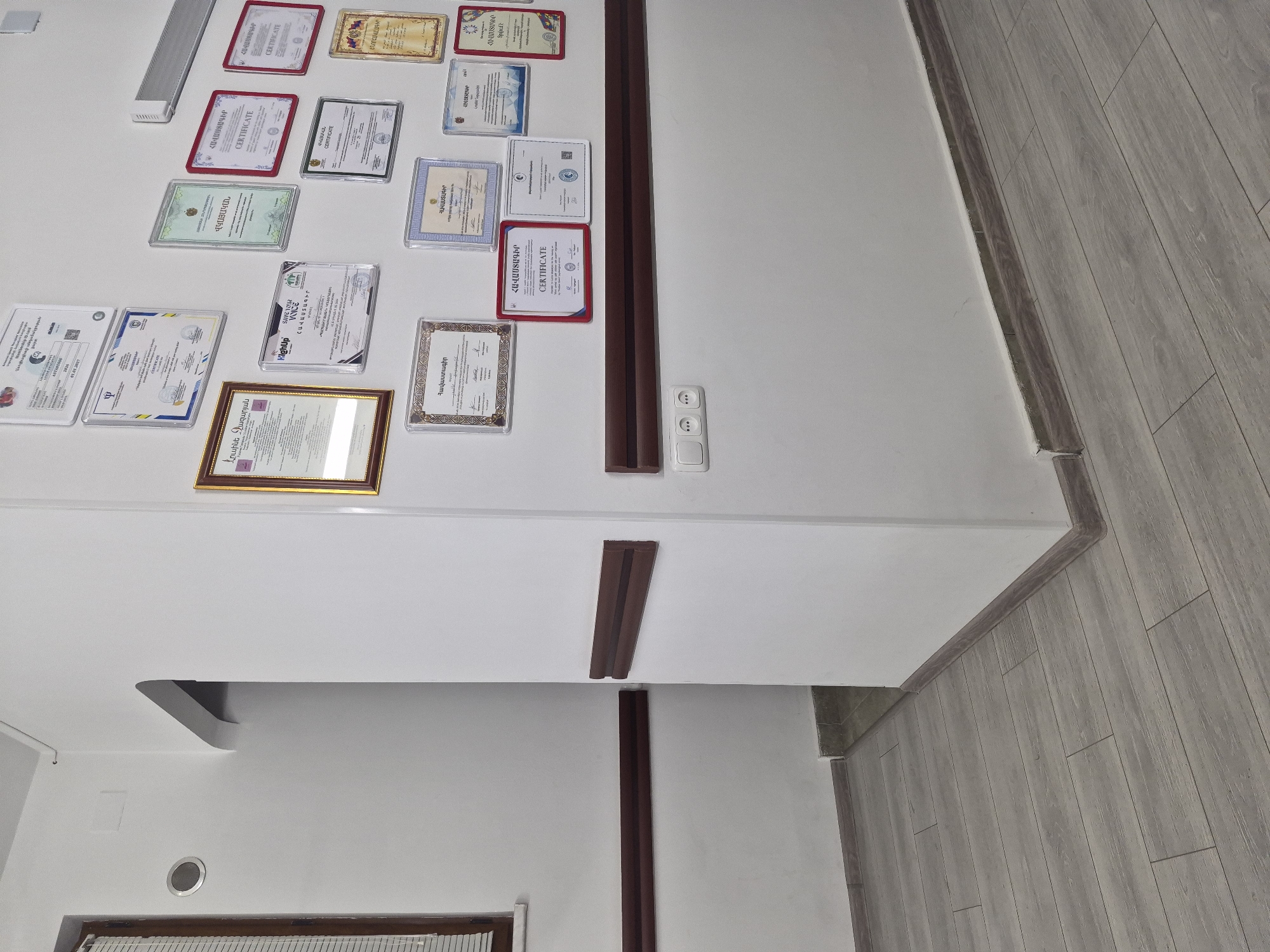 The image shows a hallway in an indoor environment, likely a professional office or institution, with a view into another room through an archway. The walls are painted white, and a dark wood-colored chair rail runs horizontally along the walls. The floor is covered with light gray wooden planks.
On the right wall, numerous framed certificates and documents are displayed at various heights. These appear to be official documents, awards, or educational certifications, predominantly in Armenian, but with some English text like "CERTIFICATE". The certificates are housed in frames of various colors, including gold, silver, and red.
In the lower part of the right wall, there is a white electrical outlet with three sockets.
The hallway area on the left shows a window with horizontal blinds, a circular vent or speaker, and a narrow doorway leading to another space. The doorway is framed by white walls and a darker, possibly tiled, threshold.
The overall impression is of a well-maintained, professional space, with a notable display of credentials. The presence of Armenian text strongly suggests a location within Armenia, and given the context of such displays, it is plausible this hallway is located in Yerevan, the capital city, in a place such as a medical facility, educational institution, or government office. However, without further identifying markers, pinpointing the exact location in Yerevan is not possible from the visual information alone.