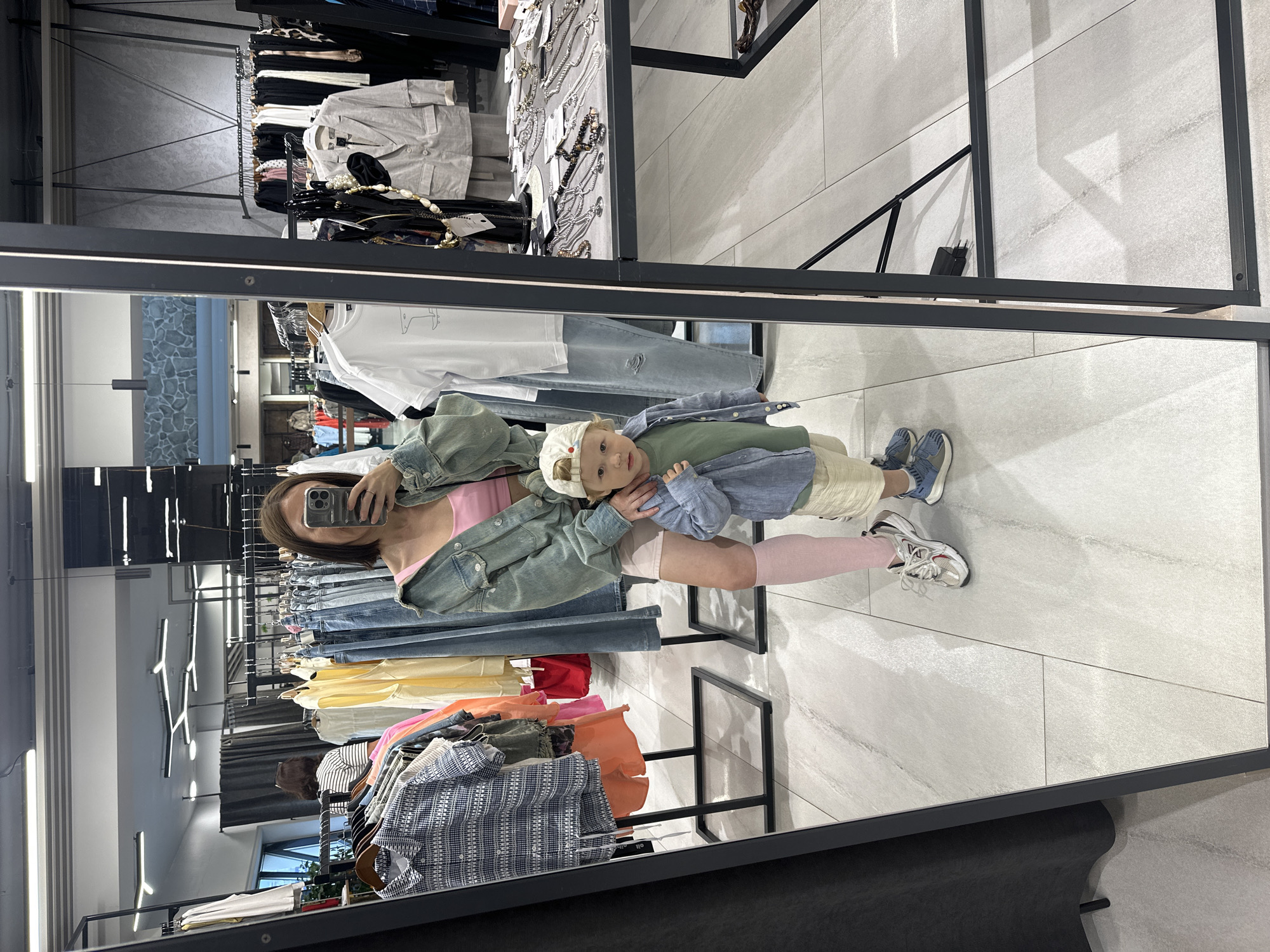The image shows a woman and a young child posing for a mirror selfie in a clothing store. The woman is wearing a pink top, a denim jacket, and pink tights. The child is wearing a green shirt, a blue button-up shirt, beige shorts, and a white cap. They are surrounded by racks of clothes and displays of jewelry, suggesting an indoor retail environment. The floor is tiled with a light gray pattern. The lighting appears to be artificial, common in retail spaces. The overall mood seems casual and perhaps playful, as they are taking a selfie together. There is no visible text in the image.