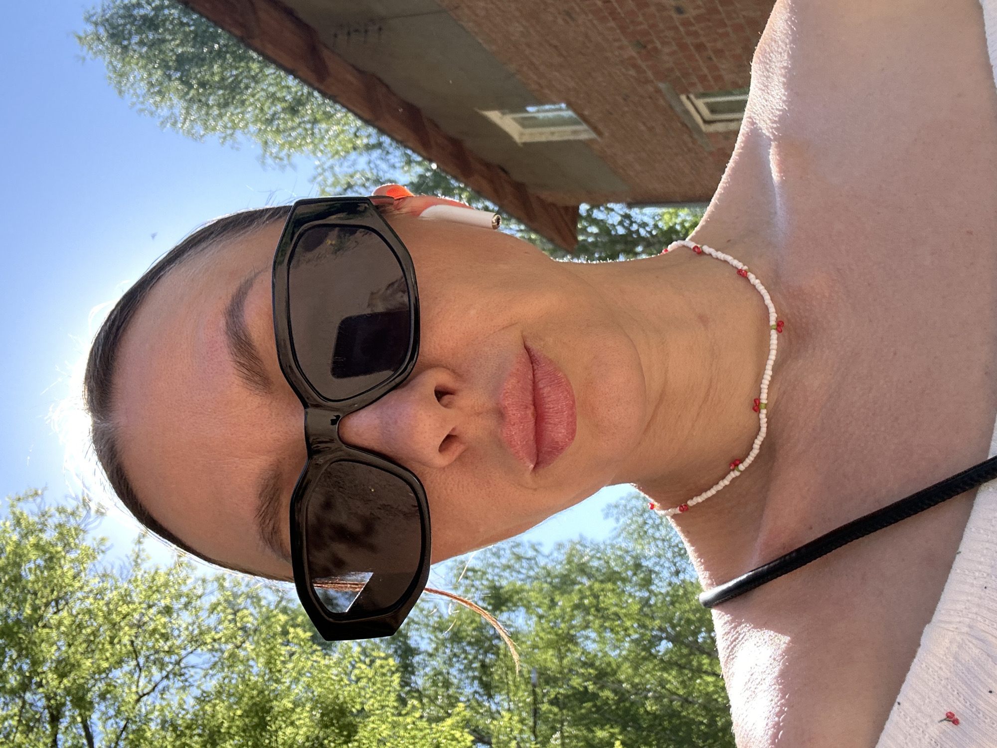 A woman with sunglasses and an earbud takes a selfie outdoors. She's wearing a beaded necklace. Behind her is a brick building, trees, and a blue sky. The location is likely in Votkinsk, Russia based on the prompt.