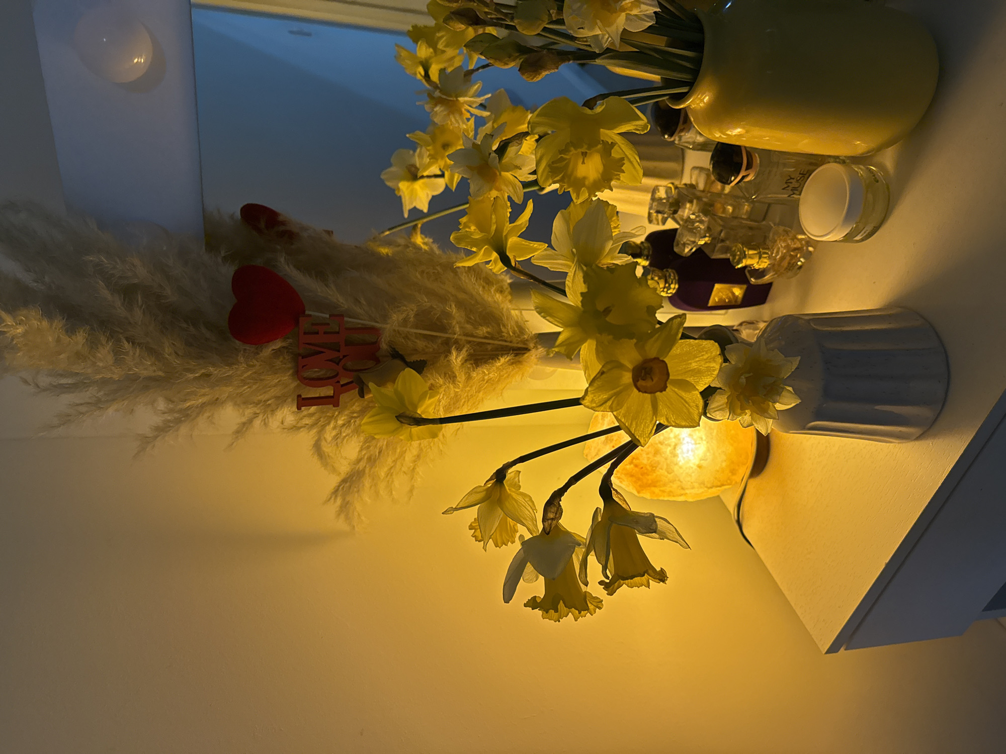 This image depicts an indoor scene, consistent with a private residence, located in Great Yarmouth, United Kingdom. The scene is presented as a still life arrangement on a white dresser top or vanity surface, viewed from a slightly elevated, frontal perspective.
On the left, an illuminated Himalayan salt lamp emits a warm, amber glow, casting yellow light across the adjacent wall and objects. To its right, a small, white, fluted ceramic vase holds several yellow daffodils. Directly in front of the lamp and extending upwards is a large arrangement of cream-colored pampas grass, within which a red decorative sign spelling "LOVE YOU" and a red heart on a stick are nestled. A second, smaller red heart is partially visible.
Further to the right, on the white surface, is an assortment of glass perfume bottles, including an opaque dark purple bottle, several clear glass bottles of various shapes, and a small, clear glass jar with white cream and a white lid. On the far right, a large, yellow, ceramic vase contains a substantial bouquet of yellow daffodils, some fully bloomed and some still in bud. Behind this arrangement, a rectangular wall mirror reflects a light-blue area, possibly a ceiling or distant wall, with a visible light source reflecting as a bright circular highlight in the upper right quadrant of the mirror. No people or active interactions are present in the scene.