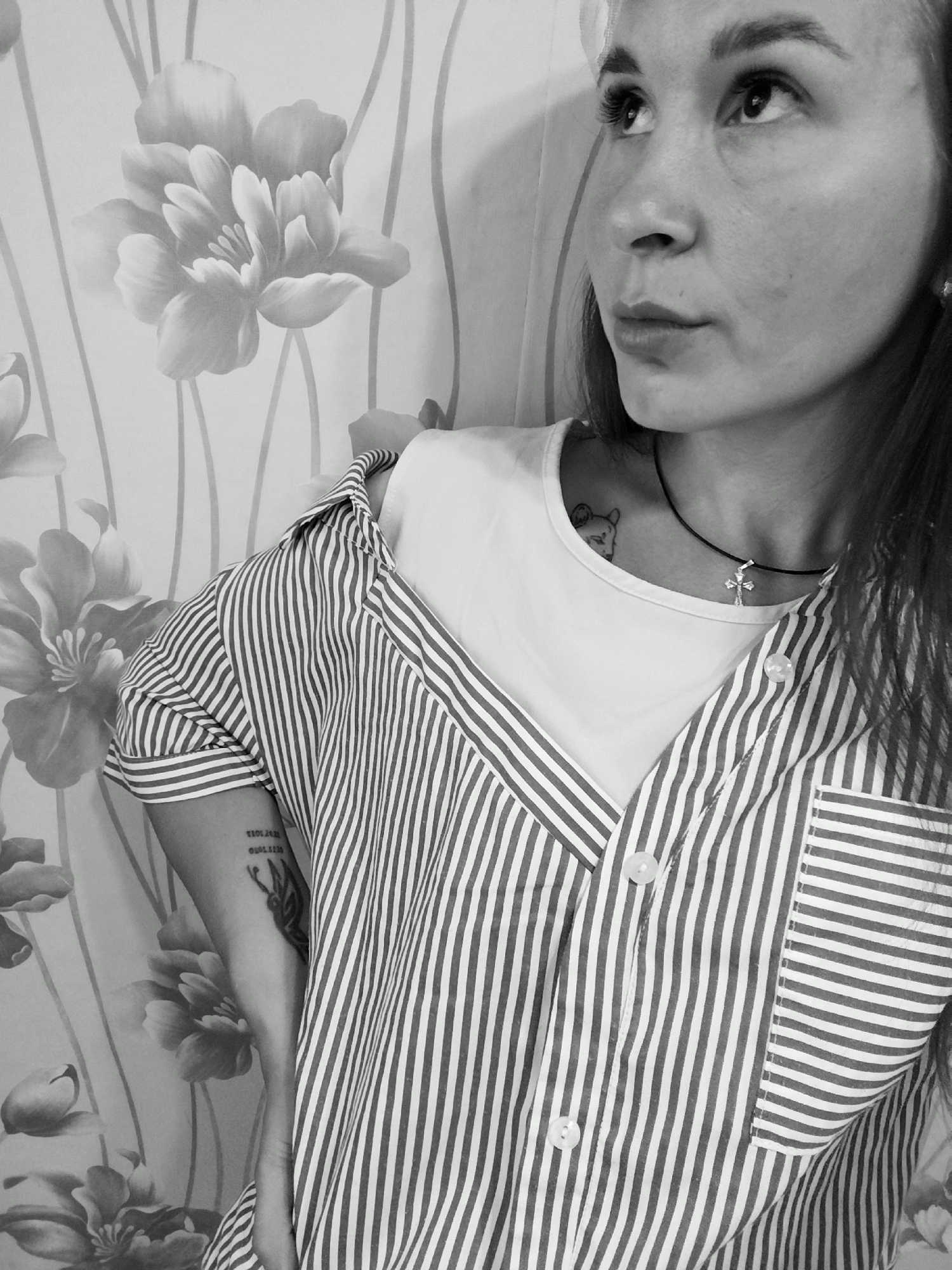 A black and white medium shot shows a young woman with long dark hair, looking up and to the right. She is wearing a white t-shirt layered under a vertically striped open button-down shirt. A thin black necklace with a cross pendant is visible around her neck. A small tattoo of a bear head is on her chest, and another tattoo of a butterfly and text is on her upper arm. She is standing in front of a floral wallpaper. The scene appears to be indoors. There is no indication of the location being within the city of Fokino, Russia.