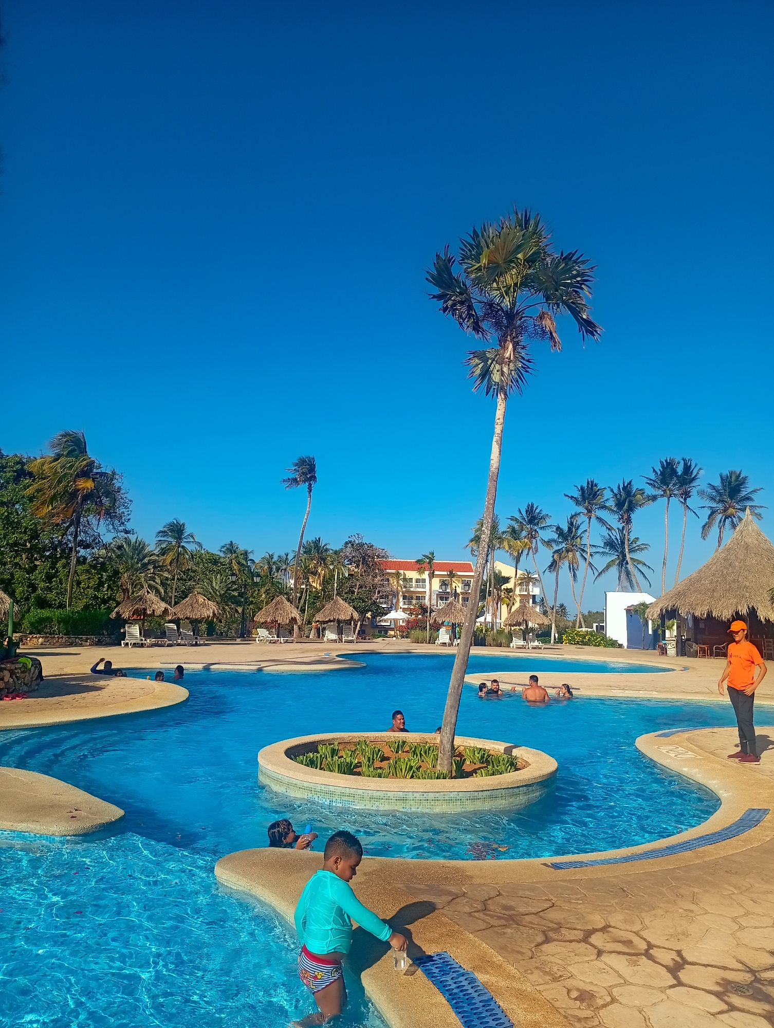 The image shows a multi-tiered outdoor swimming pool in Playa El Agua, Venezuela. The pool is surrounded by tan paving and palm trees. Several thatched-roof cabanas and lounge chairs are visible in the background. In the foreground, a young boy in a blue swim shirt and striped swim trunks stands at the edge of the pool. Several people are in the pool, including a group swimming near a small island with a palm tree. On the right side of the image, a man in an orange shirt and baseball cap stands near one of the cabanas. The sky is clear and blue.