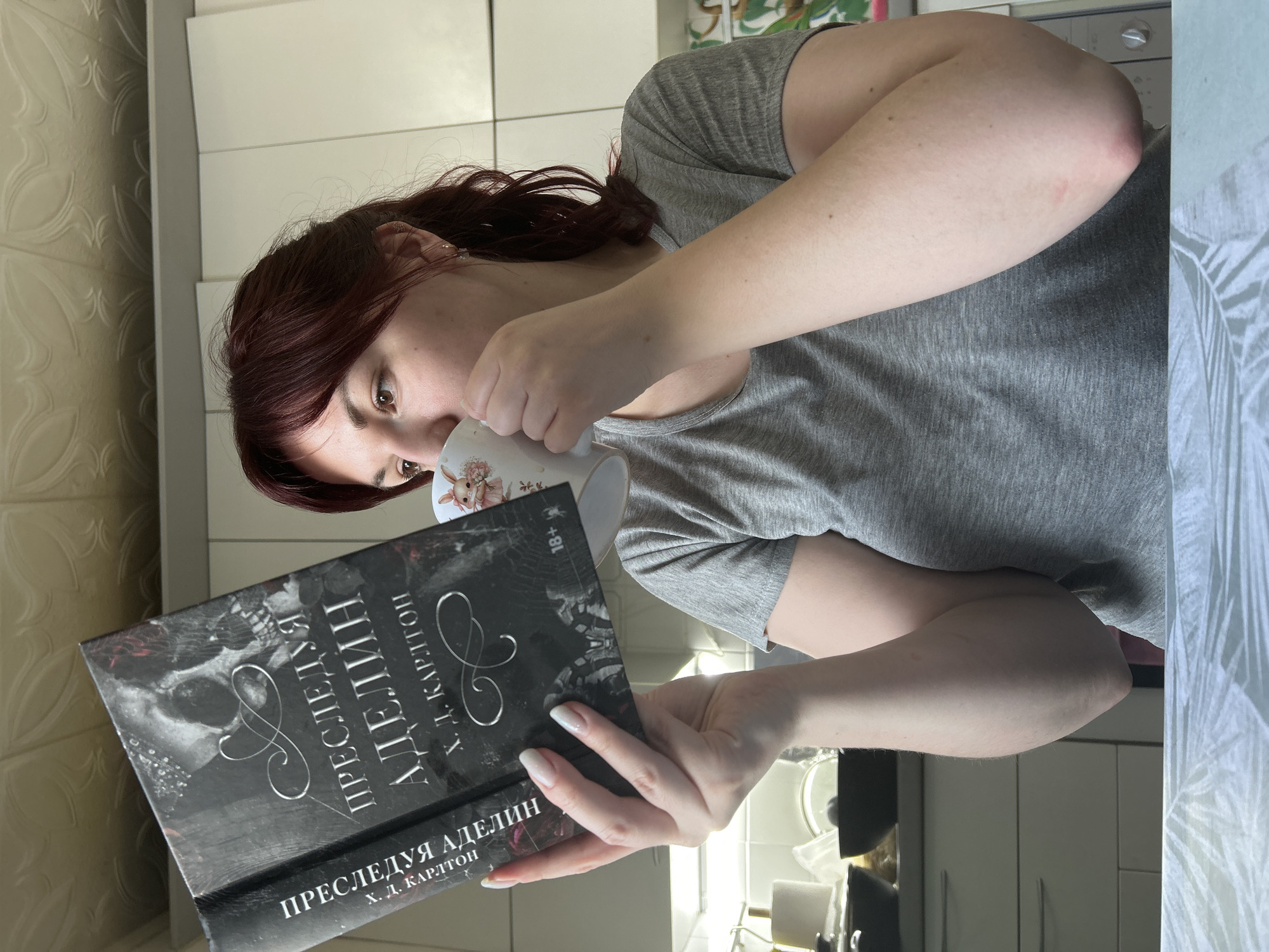 A woman with dark red hair is shown from the chest up, drinking from a white mug and holding a book in Russian. The book has a black cover with a skull-like design and the title "ПРЕСЛЕДУЯ АДЕЛИН" (Pursuing Adeline) by Х. Д. КАРЛТОН (H. D. Carlton). She is wearing a grey t-shirt. The background shows a kitchen with white cabinets. It is not possible to confirm if the location is in the city of Фоки, Russia based on the image.