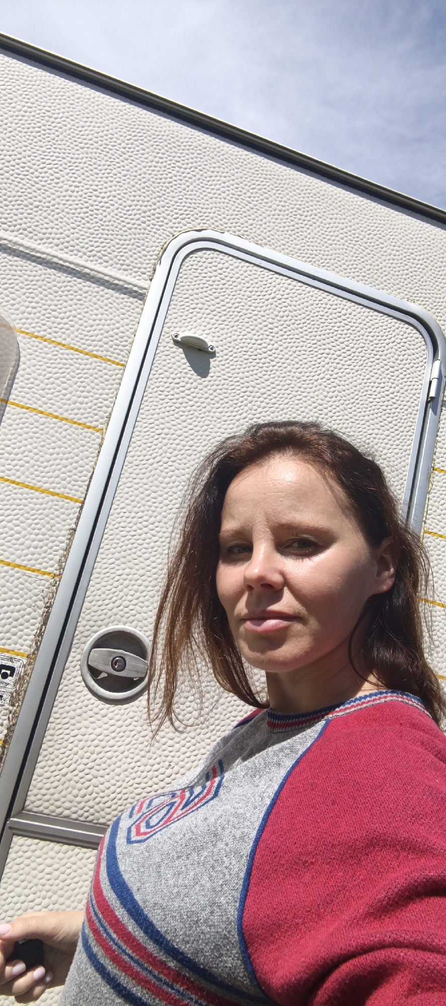 The image shows a woman taking a selfie in front of a white, textured trailer. The trailer has a silver door with a handle. The woman has brown hair and is wearing a gray and red sweater. The scene appears to be outdoors, with a blue sky visible in the background. The location is the city of Chaikovsky, Russia.