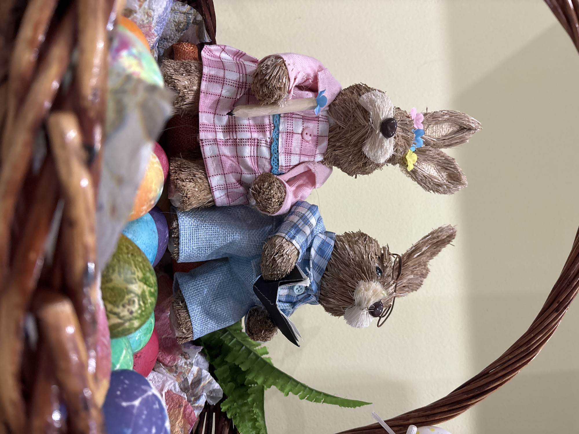 A close-up shot depicts a woven basket filled with Easter decorations. Two straw-like bunny figures are positioned side-by-side; the top bunny wears a pink and white plaid shirt and the bottom bunny wears a blue and white plaid shirt and glasses. A row of Easter eggs are lined along the basket. The background is a plain light-yellow wall. The specific location in the city of Sofia, Bulgaria, cannot be determined from the visual information provided.