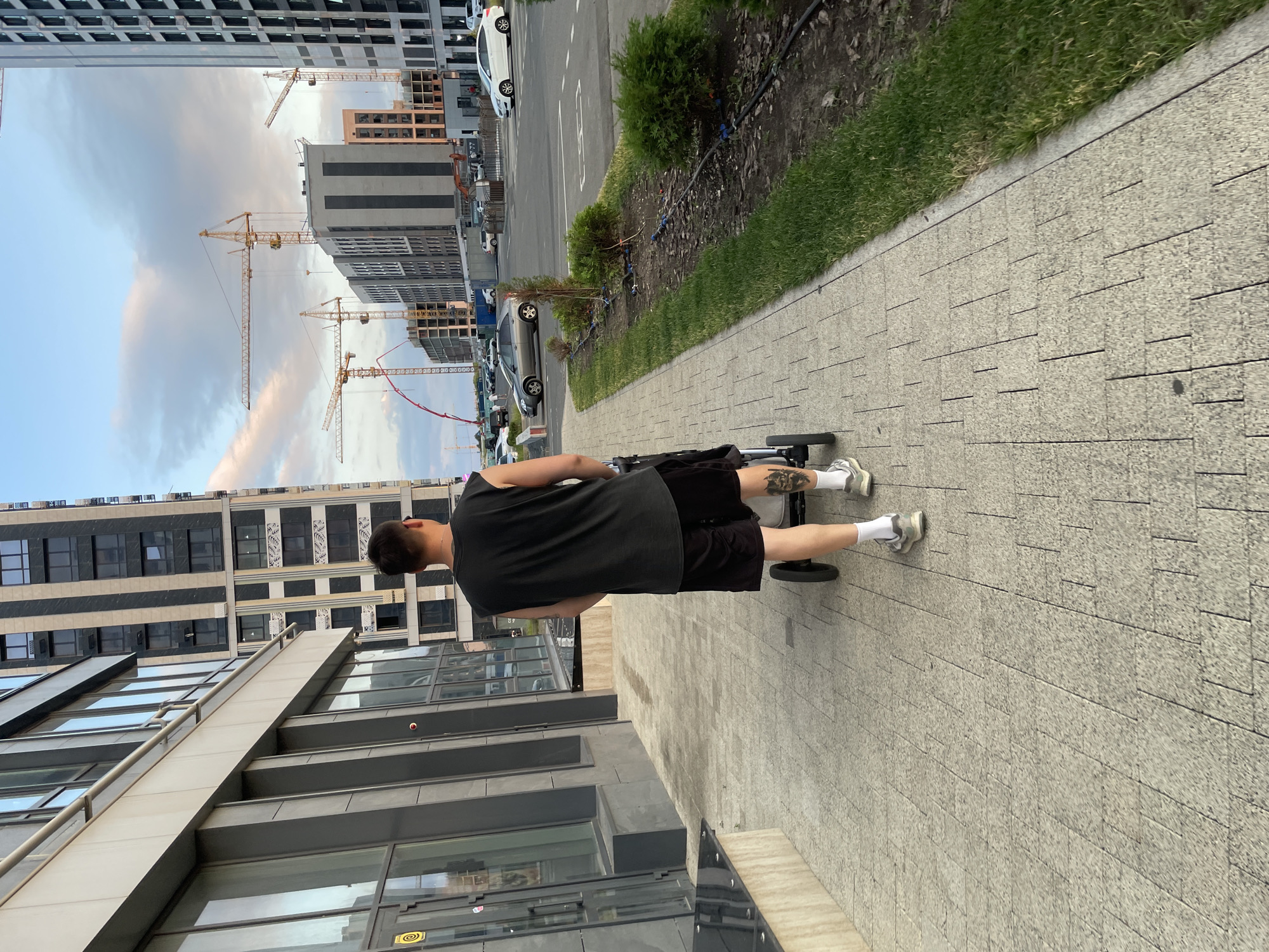 A rear view of a person walks on a paved sidewalk next to a grassy area, pushing a stroller. The person is wearing a black tank top and black shorts, with white socks and grey sneakers. A tattoo is visible on the person's left calf. To the left of the sidewalk are modern buildings with large windows. In the background, construction cranes are visible above a partially built structure and several parked cars are on a street. The sky is partly cloudy. The image is taken outdoors during the day.