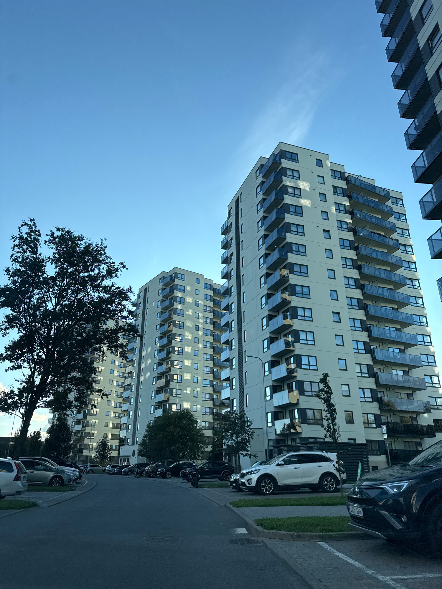 The image depicts a residential complex in Tallinn, Estonia, featuring several modern apartment buildings with light-colored facades and balconies with glass railings. A large tree with dark foliage stands prominently in the foreground to the left, partially obscuring one of the buildings. A paved road leads into a parking lot where various vehicles are parked, including a white SUV, a dark SUV, and other cars. The sky is clear and blue, with a few wispy clouds visible. The overall scene suggests a quiet, contemporary urban environment, likely during daylight hours given the illumination and shadows.