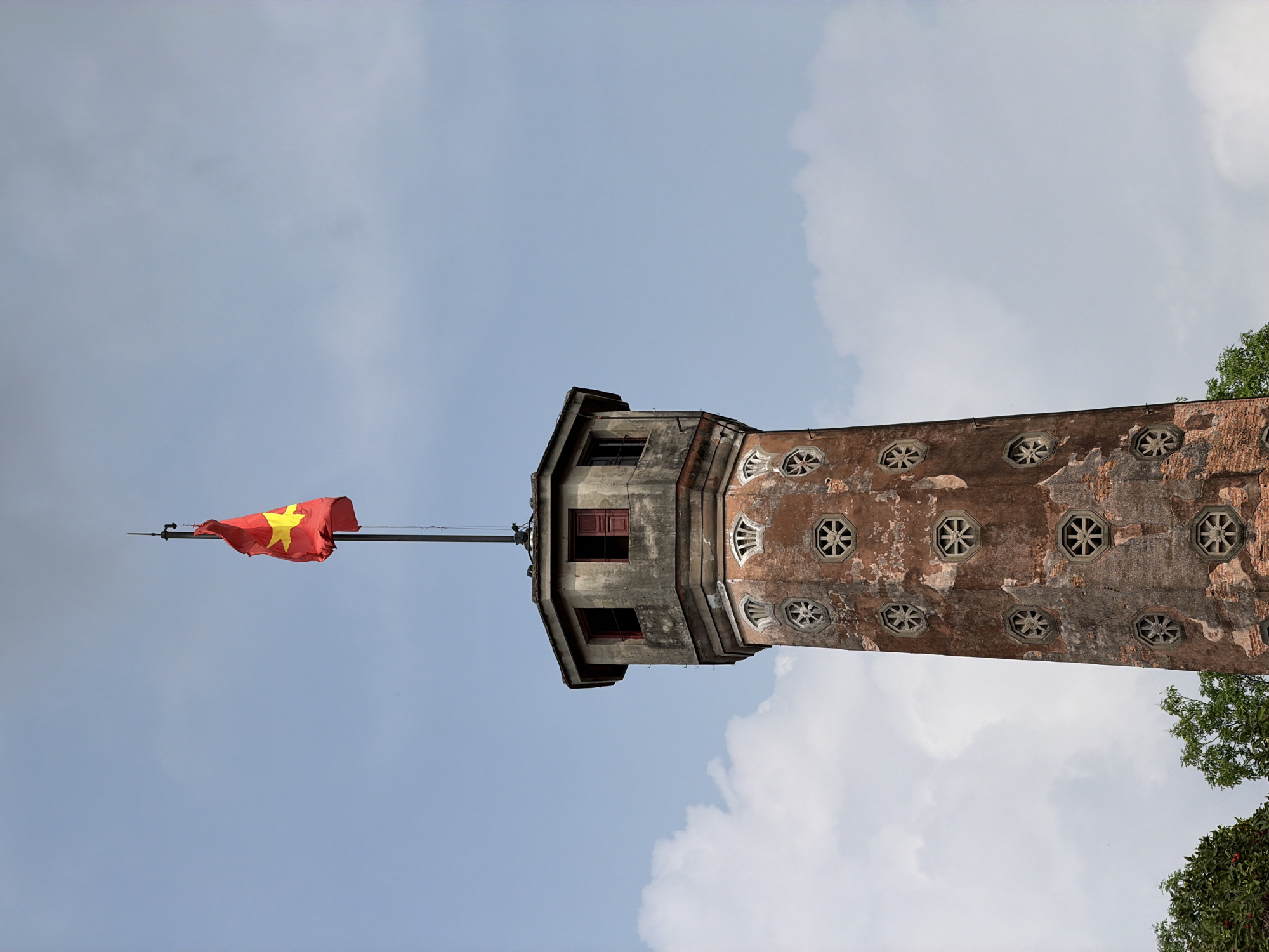 The image depicts the Flag Tower of Hanoi, located within the Hanoi Citadel, Vietnam. The tower is a tall, brick structure with a flag of Vietnam flying from its peak. The tower is set against a backdrop of a partially cloudy sky. The tower itself is composed of three levels, each level getting slightly narrower, and the overall design of the tower appears to be from a historic era. The flag is red with a yellow star. The tower is constructed of old brick and stone, suggesting age and historical significance. The tower has several round windows with decorative designs. There are trees visible on the right side of the tower.