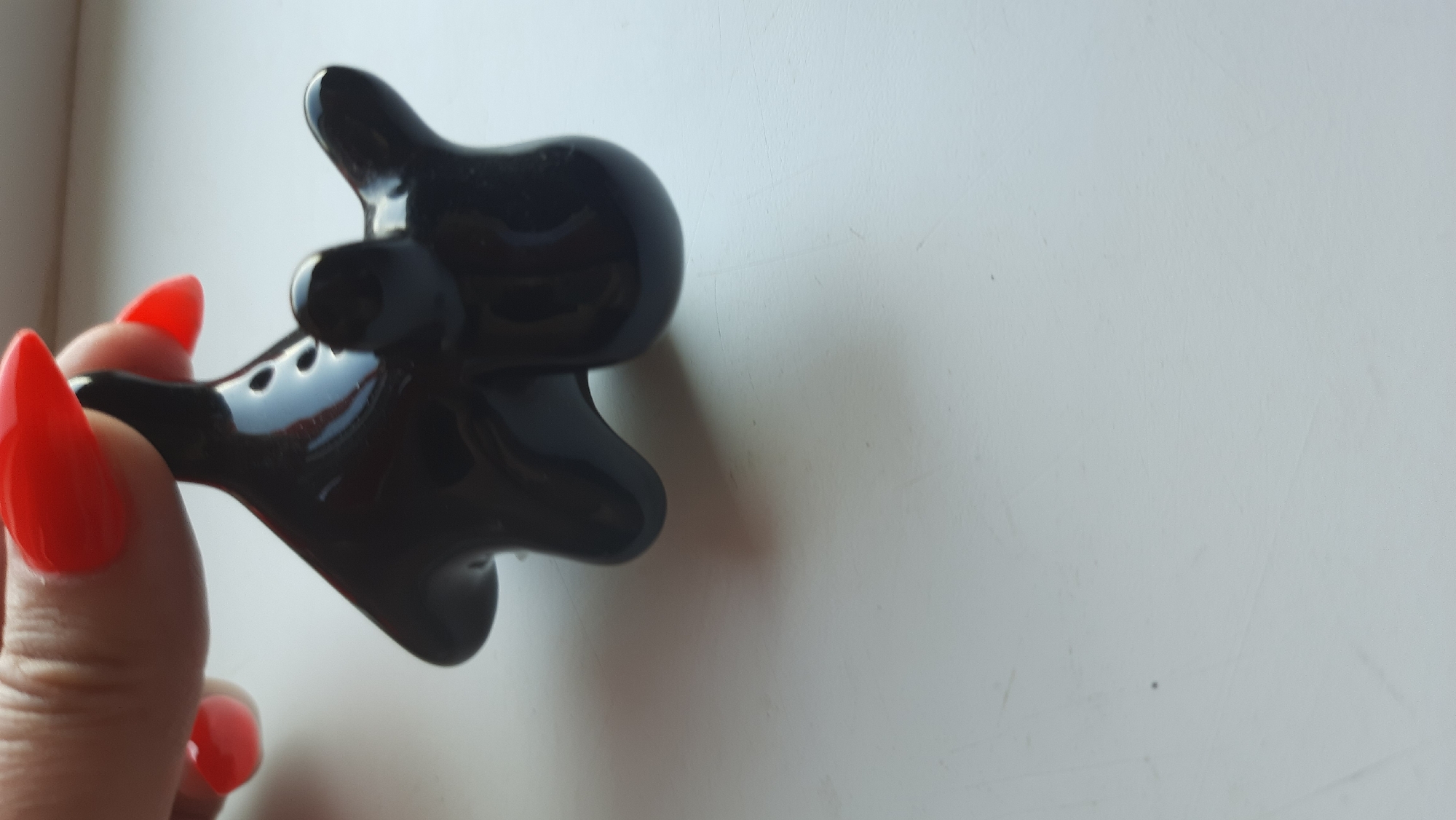 A close-up shot shows a person's hand with bright red fingernails holding a small, glossy black figurine. The figurine appears abstract and possibly made of ceramic or glass, with rounded, amorphous shapes. It is being held against a plain, white surface, likely a table or countertop, which shows some faint smudges and scratches. The lighting is bright, creating reflections on the figurine's surface. The background is out of focus, emphasizing the figurine and the hand. The scene is not identifiable as being located in the city of Desnogorsk, Russia.