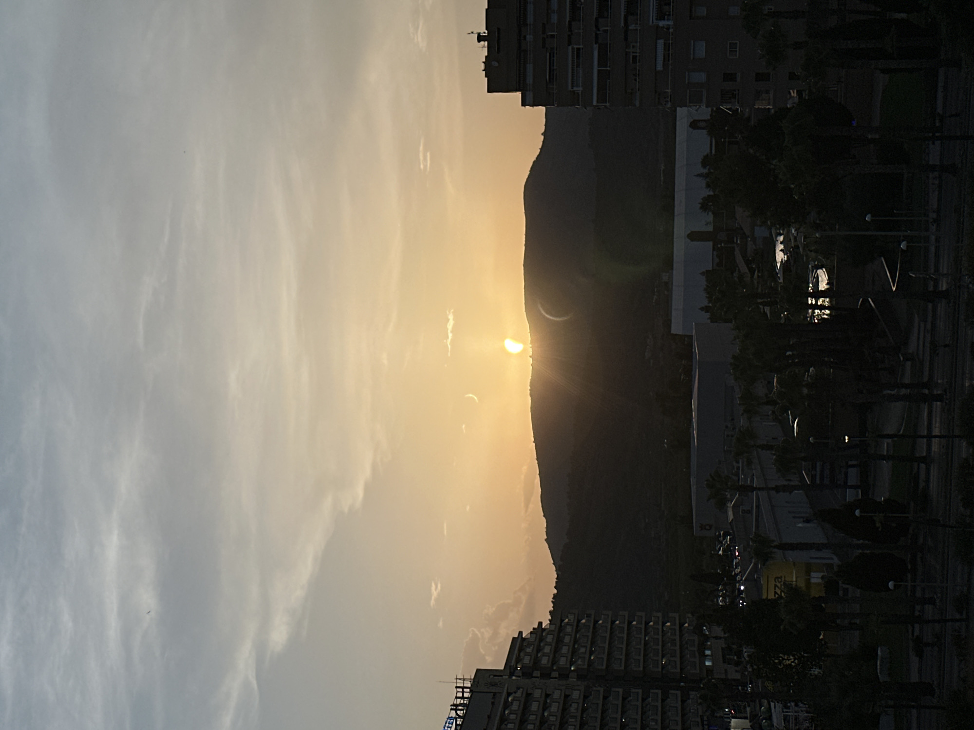 This is a vertical, medium-shot image depicting a partial solar eclipse over a silhouetted mountain range. The sun, low on the horizon, casts a warm, golden-orange glow across the sky, illuminating thin, wispy clouds. A crescent shape, the eclipsed portion of the sun, is visible against the bright orb. In the foreground, the dark outlines of buildings and palm trees suggest an urban or suburban setting. Some buildings have illuminated windows. The overall mood is serene and awe-inspiring, capturing a natural phenomenon. There is no visible text in the image. The time of day appears to be sunset or sunrise. The location is identified as Les Amplaries, Spain.