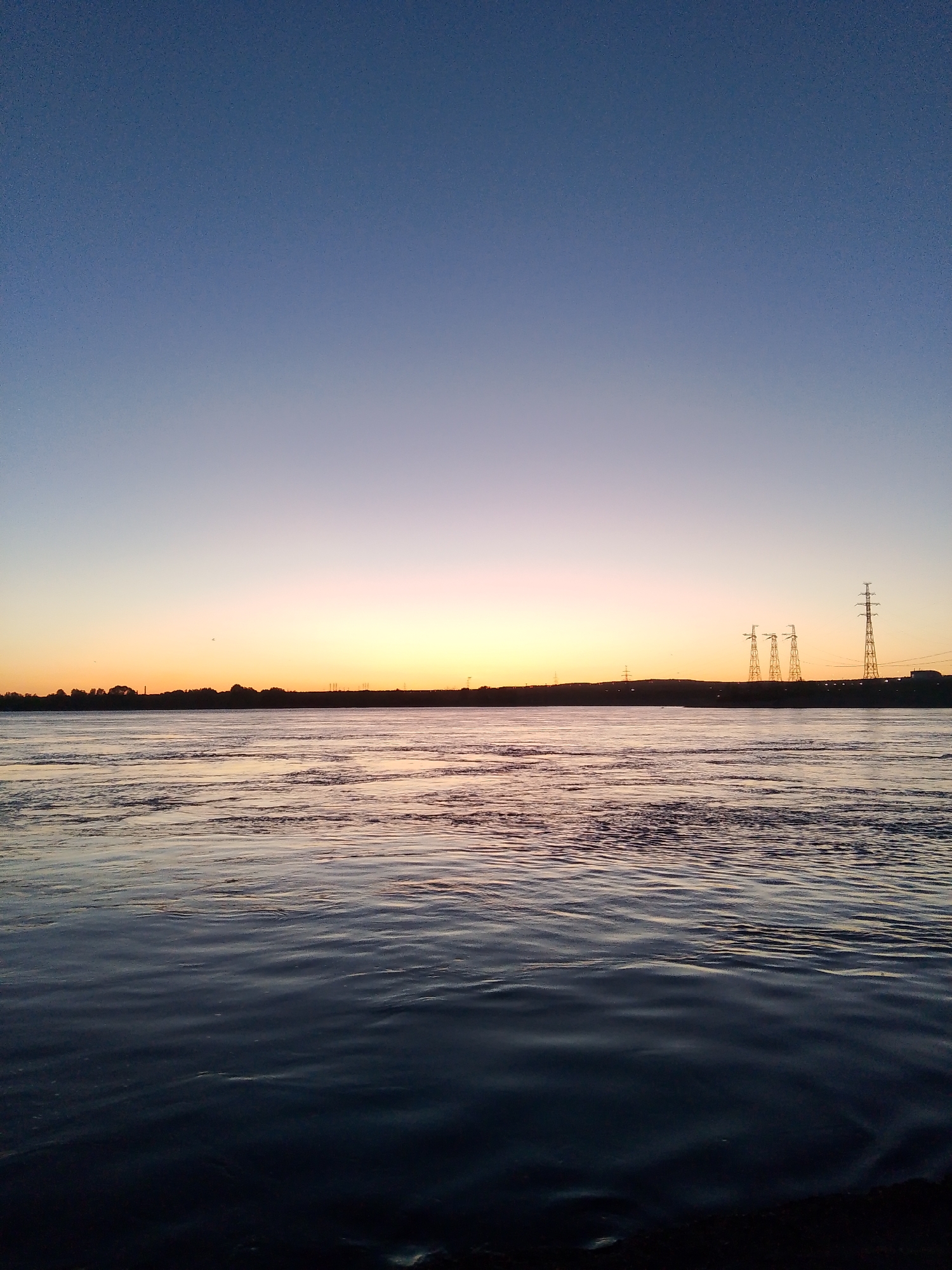 The image shows a sunset view of the Kama River in Chaikovsky, Russia. The sky transitions from a dark blue at the top to a bright orange-yellow near the horizon. The water is rippled and reflects the colors of the sky. A dark shoreline is visible at the bottom of the frame. Across the river, a silhouetted landmass is visible, punctuated by trees and power lines. There are no people visible in the image.