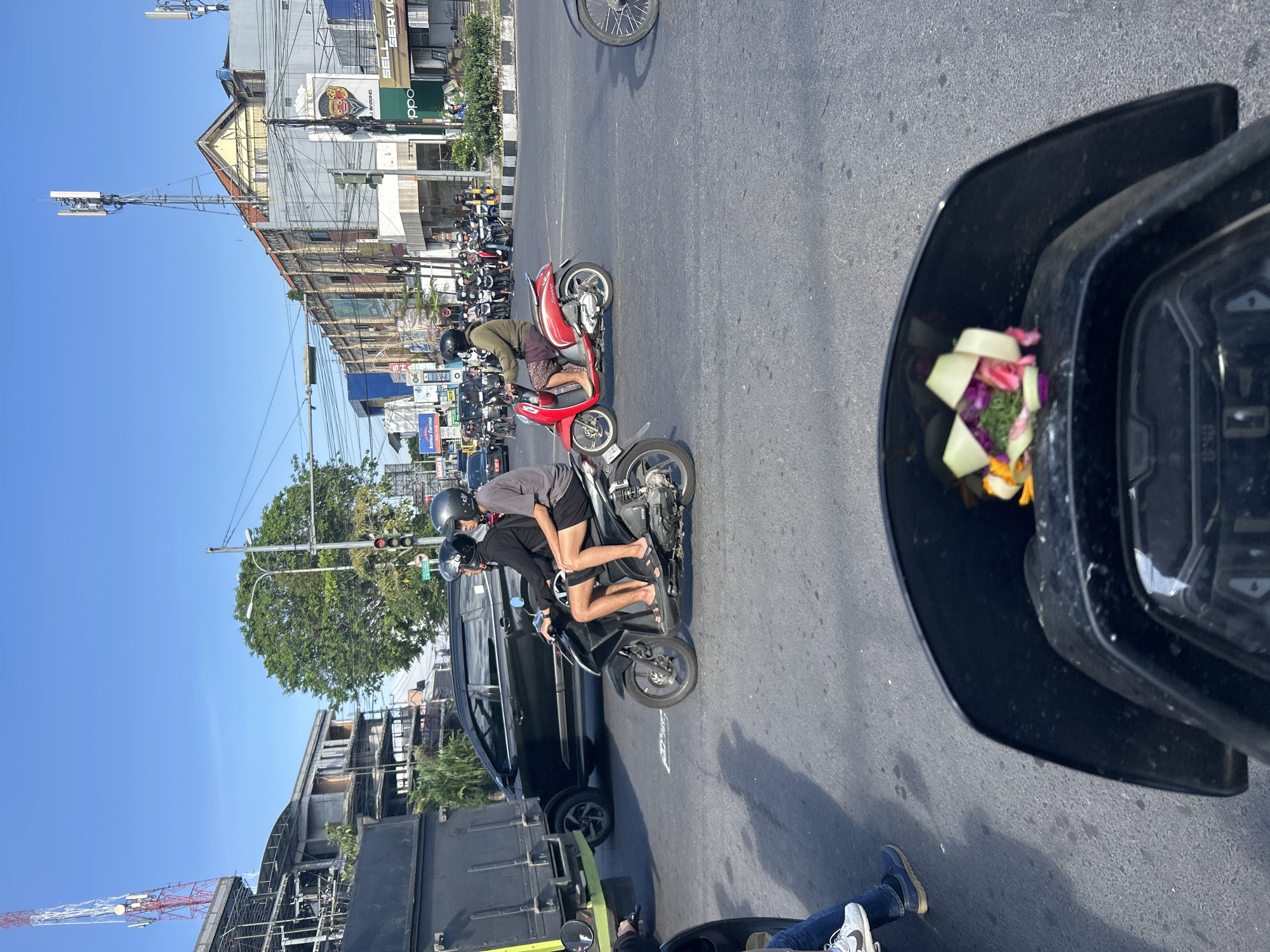 The image captures a street scene in Denpasar, Indonesia, likely during daylight hours with clear skies. In the foreground, the black dashboard and windshield of a motorcycle are visible, with a small floral offering attached to the windshield. The scene depicts traffic with several motorcycles and cars.
On the left, a black truck is partially visible. To its right, a black car is driving. Two people on a black motorcycle are in the middle ground, both wearing helmets, with the rider in front wearing a black long-sleeved shirt and shorts, and the passenger behind wearing a gray t-shirt and shorts. They are on a paved road with white lane markings.
Further to the right, a person in a green jacket and shorts is riding a red scooter, also wearing a helmet. Behind them, a line of other motorcycles with riders is visible, suggesting a traffic queue. The background shows buildings lining the street, some with visible signage and architectural details, including communication towers on top of some structures. Trees and traffic lights are also present, indicating an urban environment. The overall impression is a busy city street in Indonesia, with typical traffic and urban infrastructure.