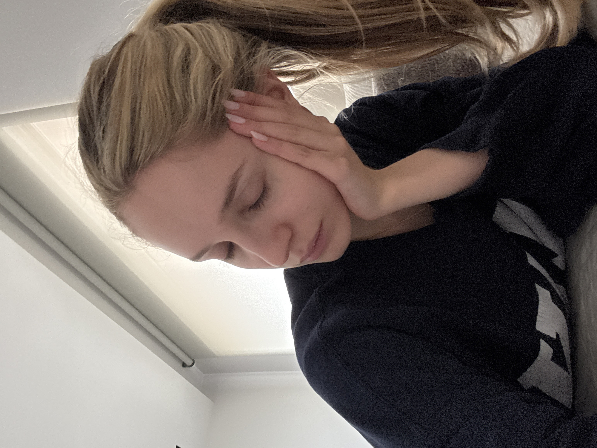 A young, fair-skinned woman with blonde hair pulled back into a high ponytail is pictured indoors, likely in a residential setting. She is wearing a dark blue sweatshirt or sweater. Her eyes are closed and her head is tilted downwards, and her hand is gently touching her face near her ear. A portion of a white window frame is visible in the background, along with a white wall. There is no immediate indication of Mexico City, Mexico, in the visual elements present. The focus is on the woman and her pose, suggesting a moment of contemplation or rest.