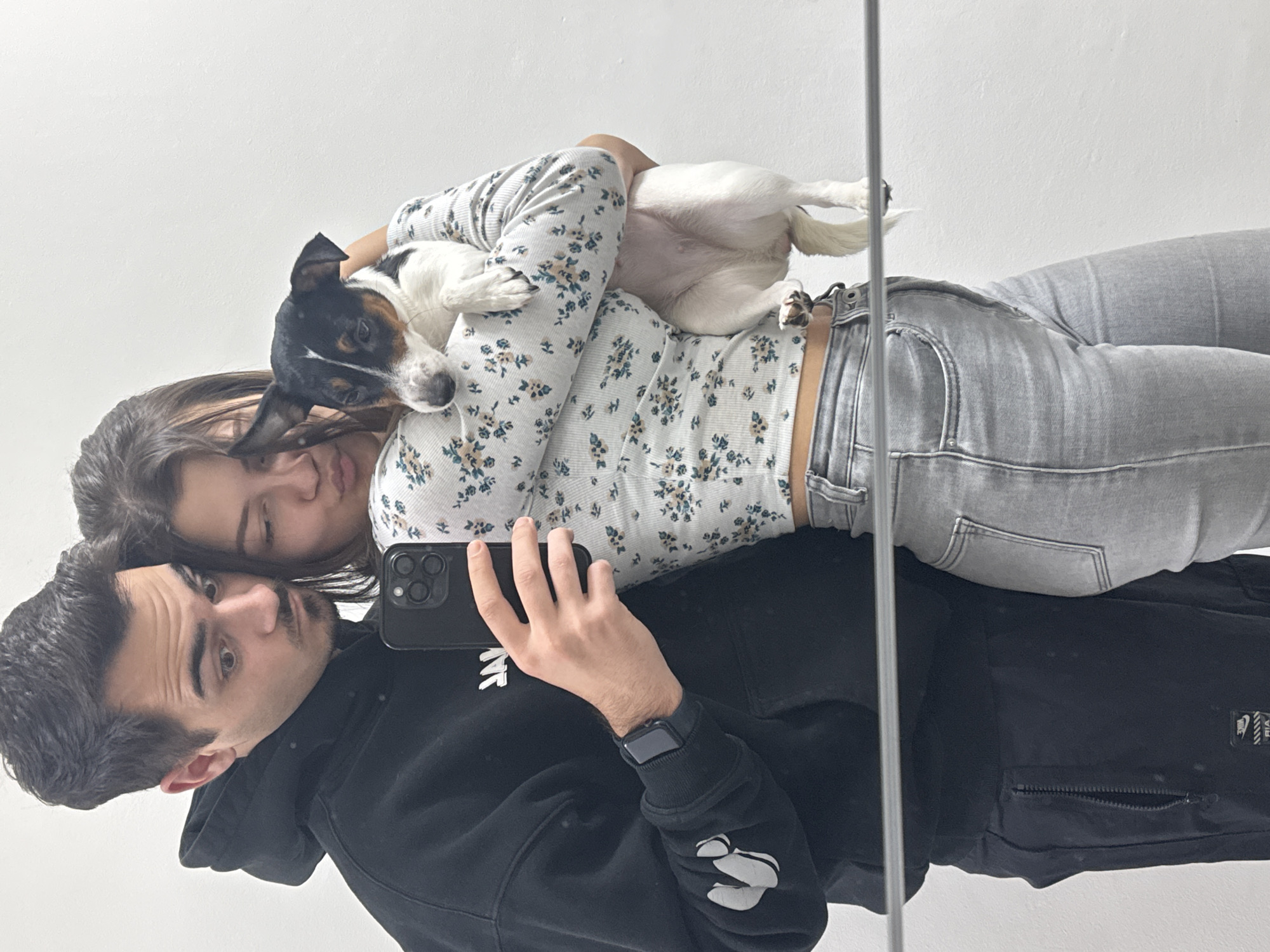 A man in a black hoodie is taking a selfie in a mirror with a woman in a floral long sleeve shirt and light gray jeans. A dog is being held by the woman. The location in Sofia, Bulgaria is not identifiable from the image.