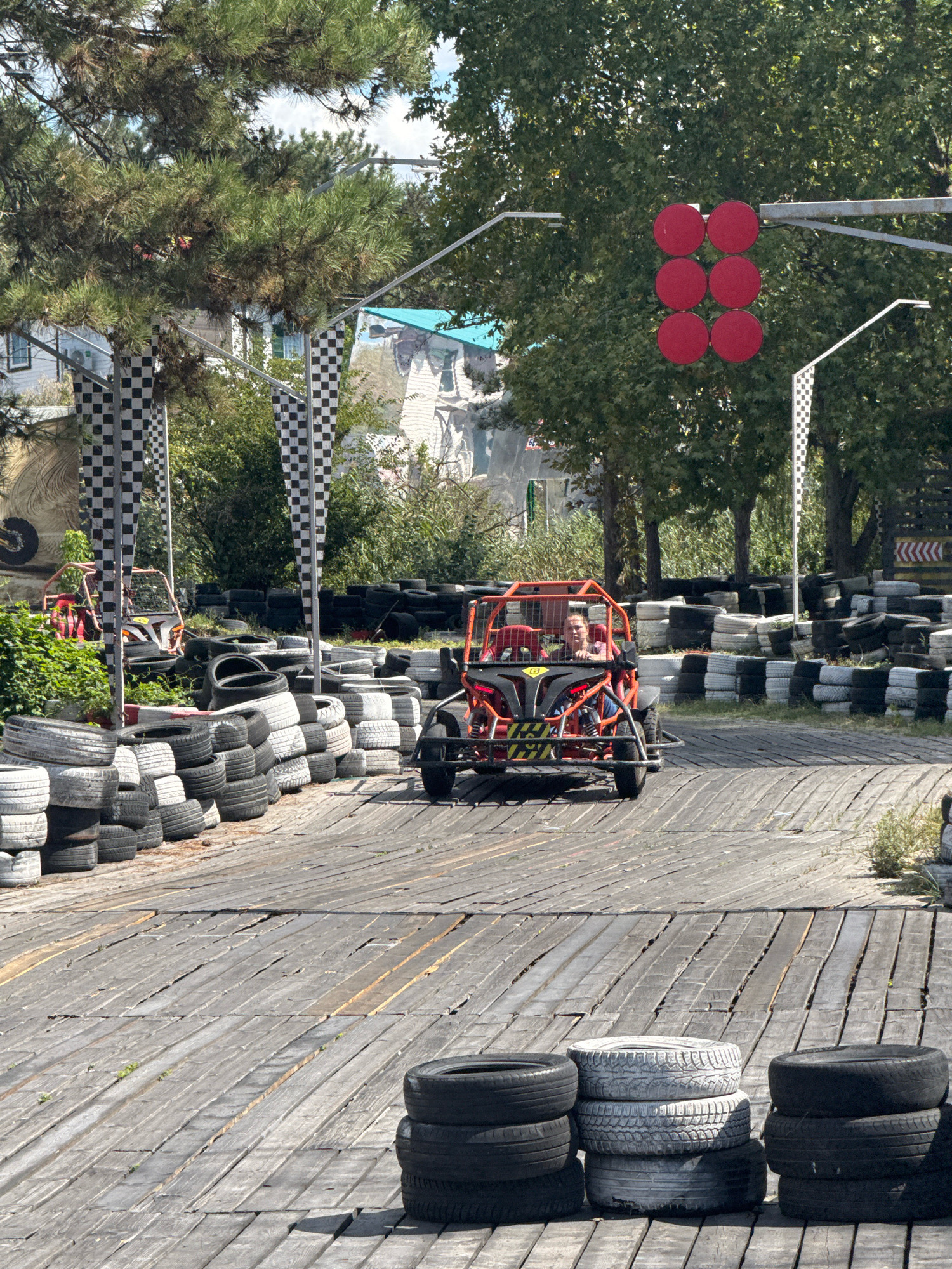 This image depicts an outdoor go-kart track, likely located in Anapa, Russia. In the foreground, stacks of used tires serve as barriers along the wooden track. A red and black go-kart is driven on the track by a person with short brown hair, wearing a dark shirt. Another go-kart, mostly obscured by tires, is parked to the right. The track is lined with more tire barriers and checkered flags on poles. In the background, lush green trees and some buildings are visible. A traffic light with six red circles is positioned above the track, indicating a signal. The sky is blue with scattered clouds. The overall scene suggests a recreational activity in a sunny outdoor environment.