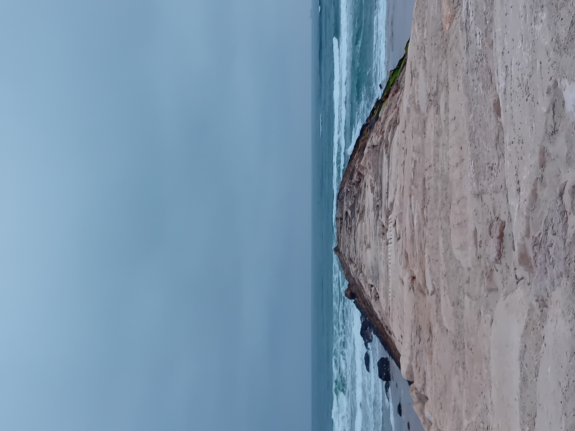 The image depicts a serene coastal landscape in Figueira da Foz, Portugal, under an overcast sky. The scene features a rugged cliff face meeting the ocean.
On the right side of the frame, a prominent, light brown or tan cliff dominates, displaying visible layers and textures indicative of sandy rock and erosion. Patches of green vegetation are visible along the cliff's edge, particularly where it meets the water.
The ocean occupies the central and left portions of the image, appearing a muted blue-gray. White-capped waves break consistently against the shore and the base of the cliff, where several dark rocks are scattered.
The sky, filling the upper-left area, is a uniform, light blue-gray, suggesting an overcast or hazy daytime. The light is diffused, with no direct sunlight.
No people or identifiable man-made structures are visible. The overall atmosphere is one of natural ruggedness and tranquility. There is no visible text in the image.