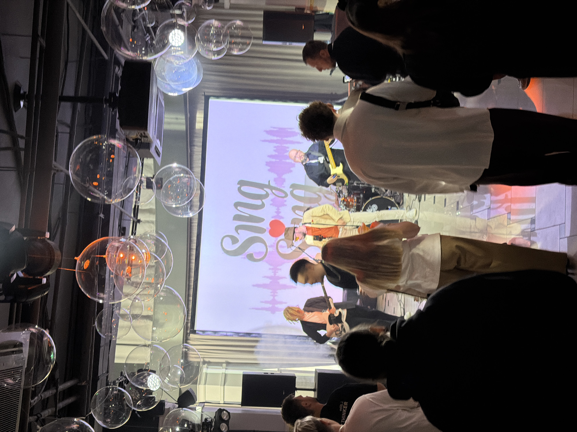 A gathering of people is occurring, seemingly at a venue in Perm, Russia. A band is performing on stage in front of a projected screen displaying a design that includes the word "Sing." Some in the audience are facing the stage, presumably watching the performance, while others are engaged in other activities. Overhead are decorations consisting of numerous clear, spherical objects, possibly balloons or light fixtures. The lighting in the venue is dim, creating a relaxed and social atmosphere. The scene also includes architectural elements, equipment related to the band's performance, and other event-related accessories.