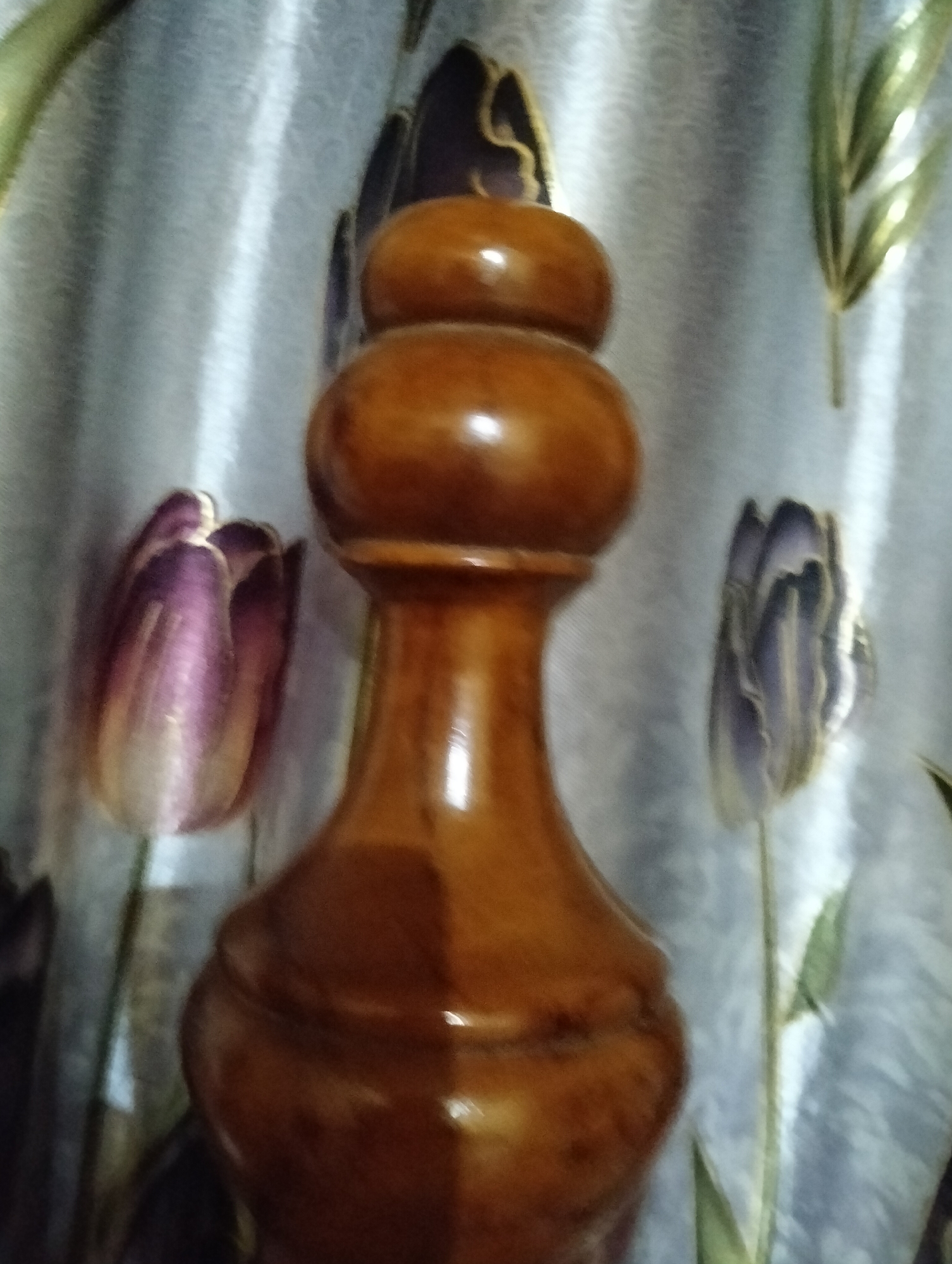 The image shows a wooden finial, possibly from a piece of furniture, against a background of floral patterned curtains. The finial is brown and has a smooth, polished finish with a rounded, decorative top. The curtains are light-colored with a pattern of purple and green tulips and leaves. The scene is indoors, likely in a bedroom or living room. There are no people or animals visible in the image. The location in the city of Payagaboh, Indonesia, is not specified or inferable from the visual information.