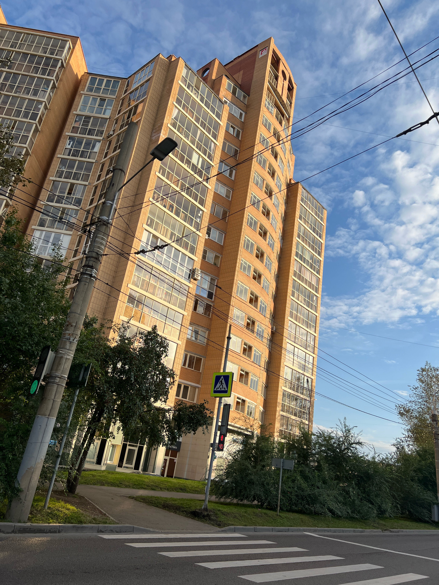 The image captures a view of a modern, multi-story residential building with a beige brick facade and numerous glass balconies, set against a clear blue sky with scattered white clouds. In the foreground, a pedestrian crossing is marked on an asphalt road, and a sidewalk runs alongside a grassy area. A concrete utility pole with electrical wires, a traffic light showing green, and a street lamp are prominent on the left side of the image. A pedestrian crossing sign is visible near the building's entrance, accompanied by a traffic signal displaying red. Dense green trees and bushes line the perimeter of the building. The architecture suggests an urban setting, and the presence of a pedestrian crossing and traffic signals indicates a public street. This scene is located in the city of Krasnoyarsk, Russia.