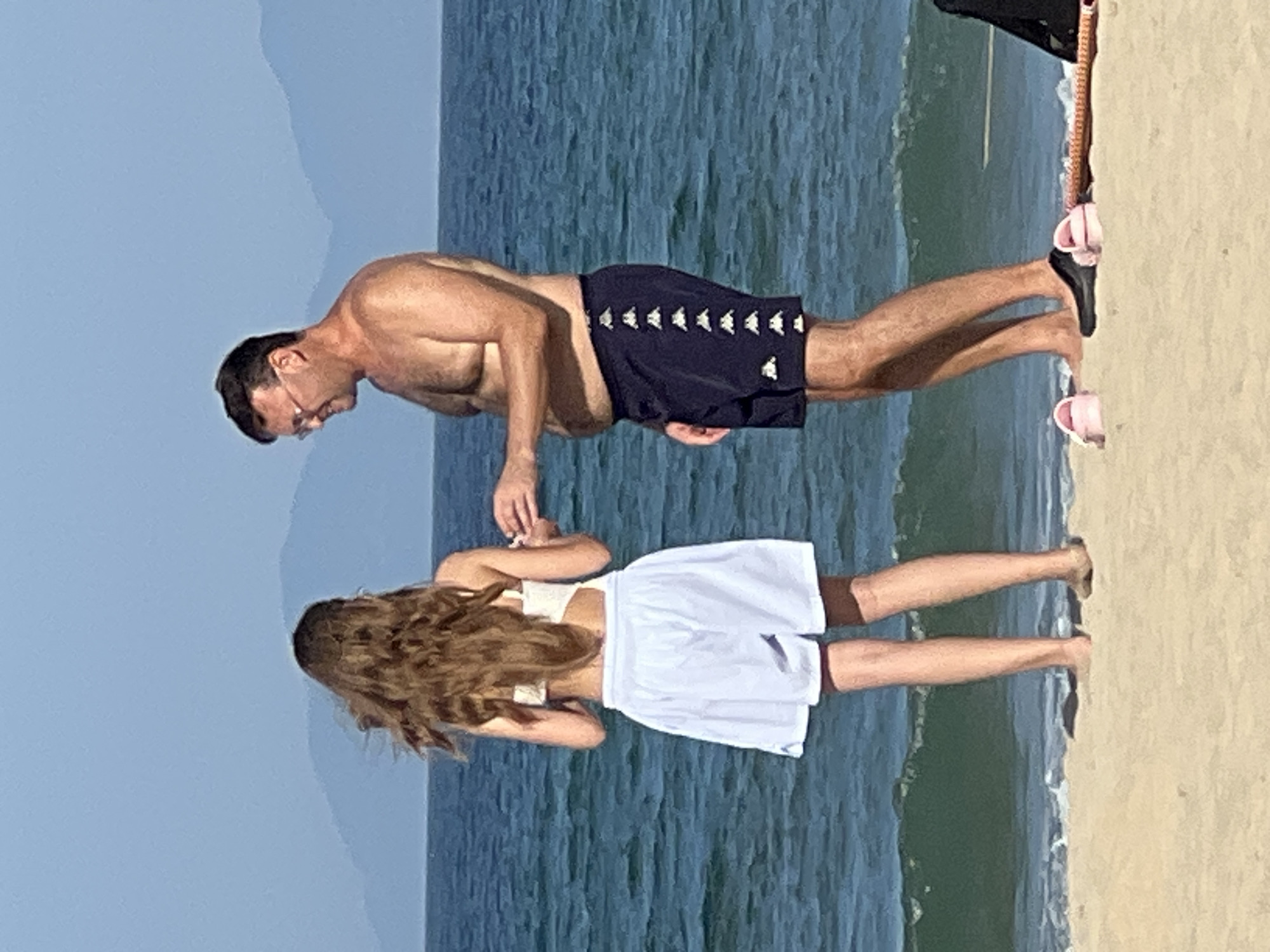 Two people stand on a sandy beach in Da Nang, Vietnam, with the ocean in the background. A woman with long, brown hair, wearing a white bikini top and white shorts, faces away from the camera. A man, wearing dark swim trunks with white accents, stands to her right, facing her. He has short brown hair and is wearing glasses. They appear to be interacting, with the man's right hand extended towards the woman. The ocean is a deep blue with moderate waves, and distant mountains are visible on the horizon under a clear blue sky. The sand in the foreground shows footprints and a few scattered flip-flops.