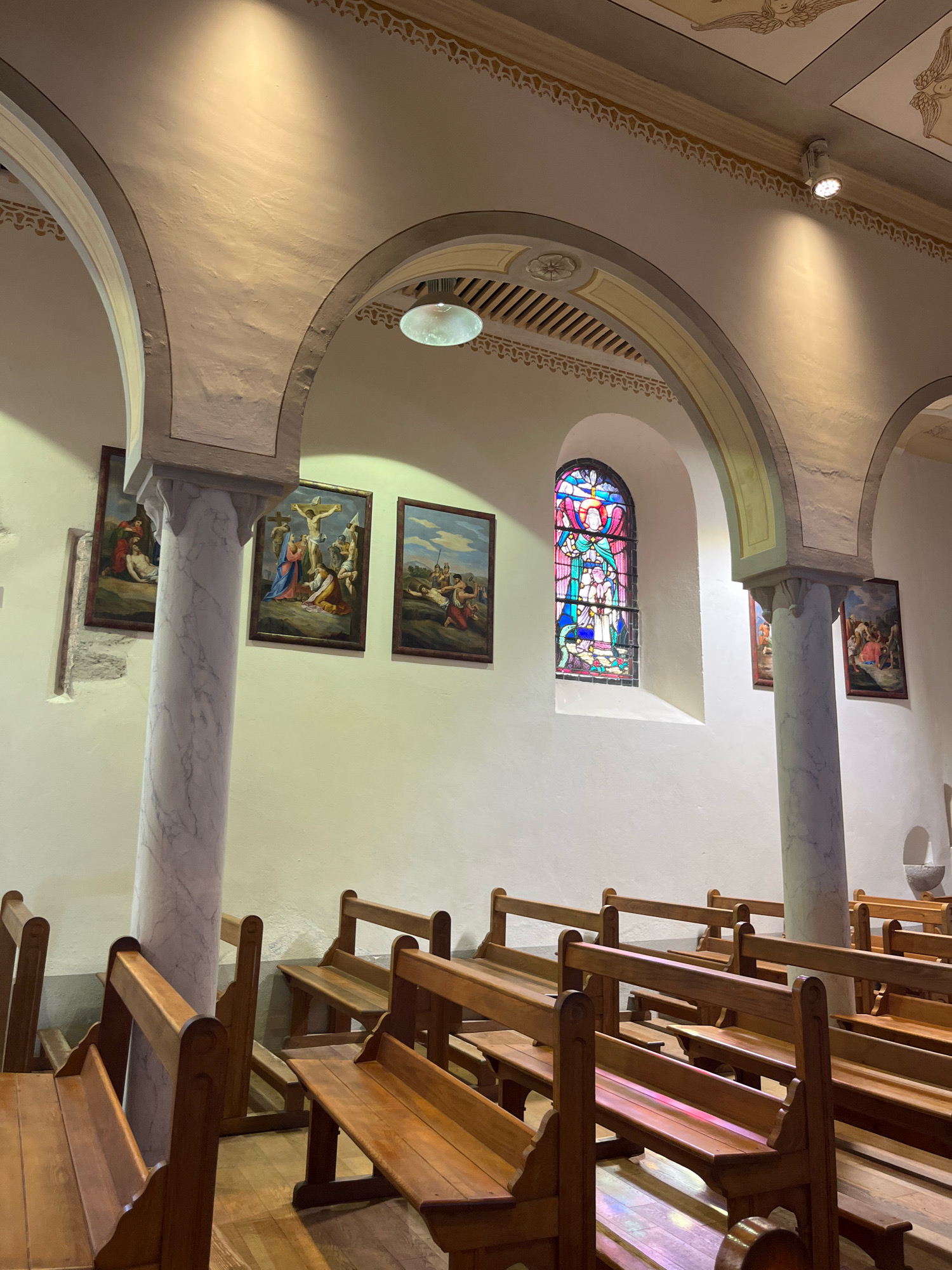 The image depicts the interior of a church in Yvoire, France. The scene is dominated by rows of empty wooden pews facing a central aisle. The walls are painted a light color and adorned with religious paintings depicting scenes from the life of Jesus Christ, including the crucifixion. To the right, a stained-glass window with colorful geometric and figural patterns is visible. The ceiling features arched structures and decorative moldings, with lighting fixtures casting warm light on the surfaces. The floor is made of polished wood. There are no people present in the image. The overall atmosphere is one of solemnity and quiet contemplation.