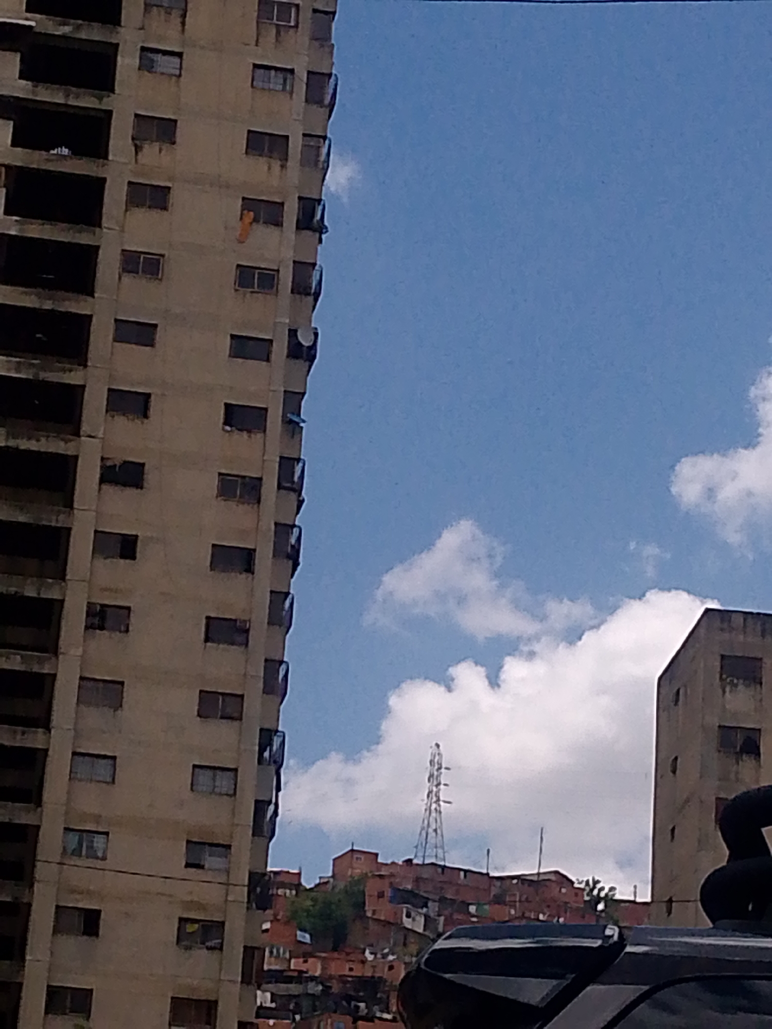 The image shows a partial view of Caracas, Venezuela. Two tall, dilapidated concrete buildings dominate the scene, the one on the left more extensively so. Many window openings are empty, and the concrete is stained and weathered. Between the buildings, a densely packed hillside of small, red-brick houses rises. A tall communications tower is visible on the hill. In the foreground, part of a dark vehicle (likely a car) is visible, partially obstructing the view of the hillside. The sky is blue with scattered white clouds.