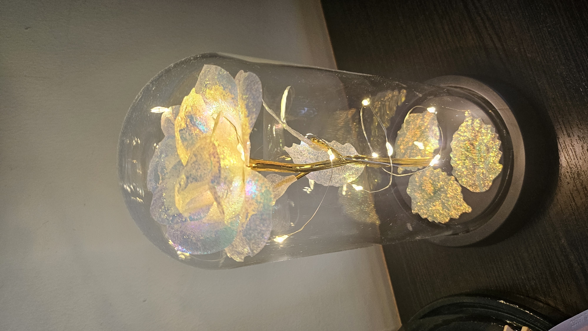 The image shows a close-up, indoor shot of a decorative object. The main subject is a faux rose, encased in a clear glass dome. The rose petals have an iridescent, pearlescent finish, reflecting light in shades of yellow, white, and hints of pink and blue. Delicate string lights with small, warm white LEDs are wound around the stem and petals of the rose, illuminating it from within. The stem and leaf elements are metallic gold. The glass dome sits on a dark, circular base. The background is divided, with the left side appearing to be a light-colored, textured wall, and the right side showing a dark, wooden surface. The lighting suggests it is not daytime, and the overall mood is decorative and ambient, possibly intended for interior decoration. No people or text are visible in the image.