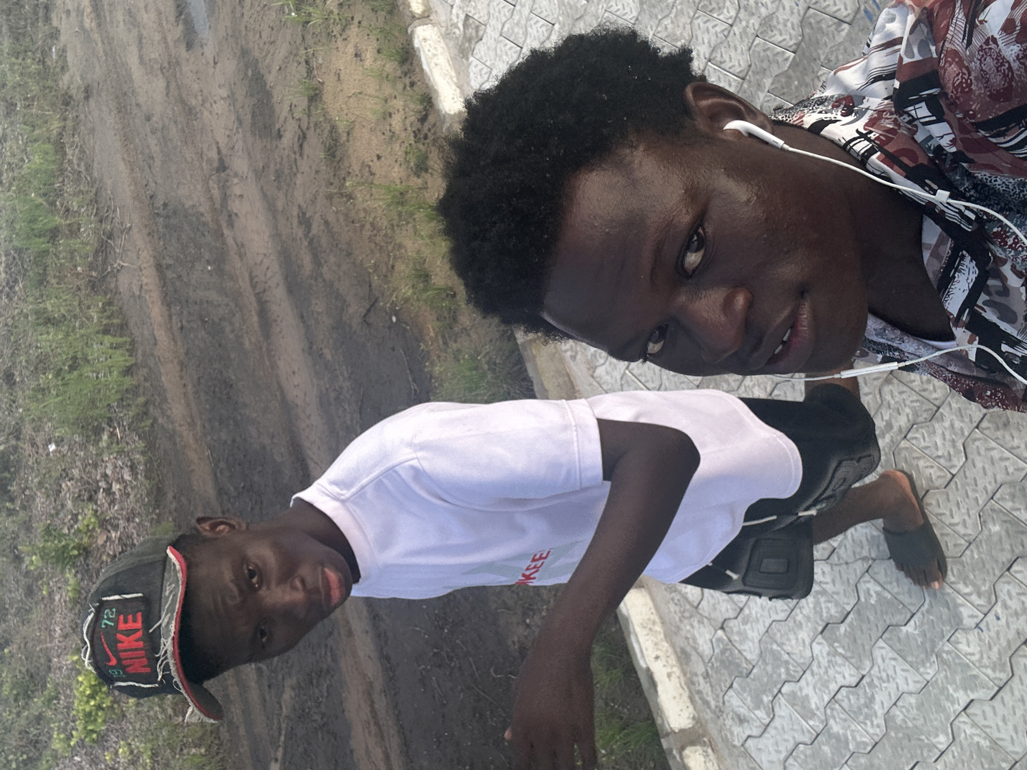Two young Black men are walking on a paved walkway in an outdoor environment. The man on the left is wearing a black Nike hat with red lettering, a white t-shirt, and black shorts. He is looking at the camera with a serious expression. The man on the right is wearing a multi-colored patterned shirt and white earbuds. He is also looking at the camera, with his face closer to the lens. Behind them, the ground is a mix of dirt and some sparse vegetation. The walkway is made of interlocking grey pavers. The overall scene appears to be in a relatively undeveloped or transitional area, potentially on the outskirts of a city.