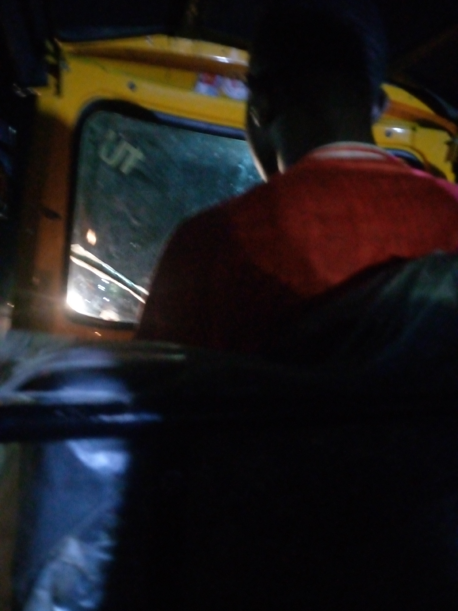 The image shows the interior of a yellow vehicle, likely a tuk-tuk or a small bus, at night in Jalingo, Nigeria. A person wearing a red shirt is visible from behind, seated in the vehicle. The person's head is silhouetted against the dark interior and a blurred view through the window. The window shows the reflection of lights, suggesting movement. The environment appears to be urban, with the characteristic nighttime lighting of a Nigerian city. The overall image is dark and somewhat grainy, indicating low light conditions during the capture.