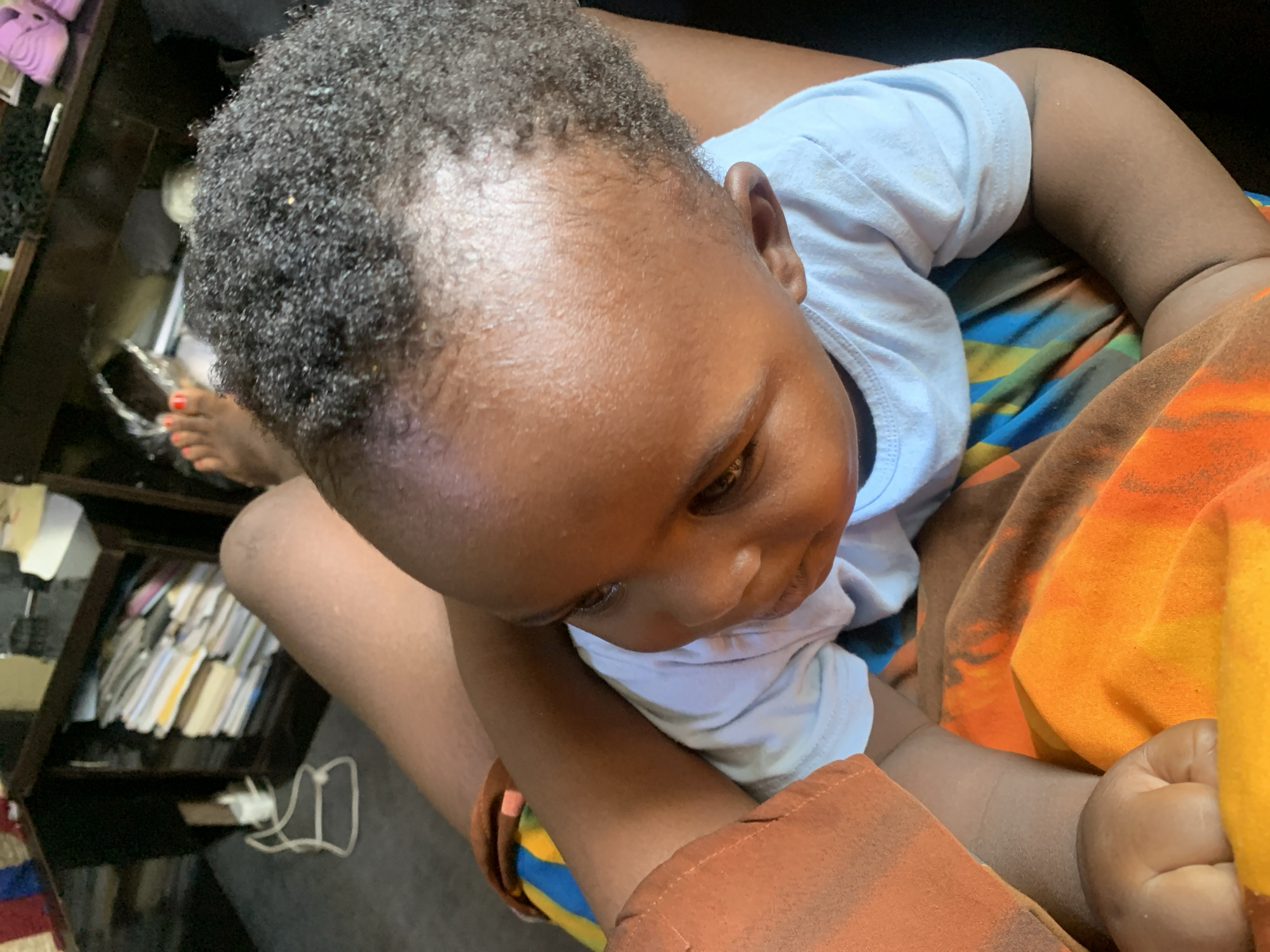 The image shows a close-up view of a baby sitting on someone's lap. The baby is wearing a light blue t-shirt and has dark, curly hair. The person's lap is covered with a colorful fabric. In the background, there is a dark wooden shelf unit filled with books and other items. There are also some pink shoes visible on the top shelf. The scene appears to be indoors.