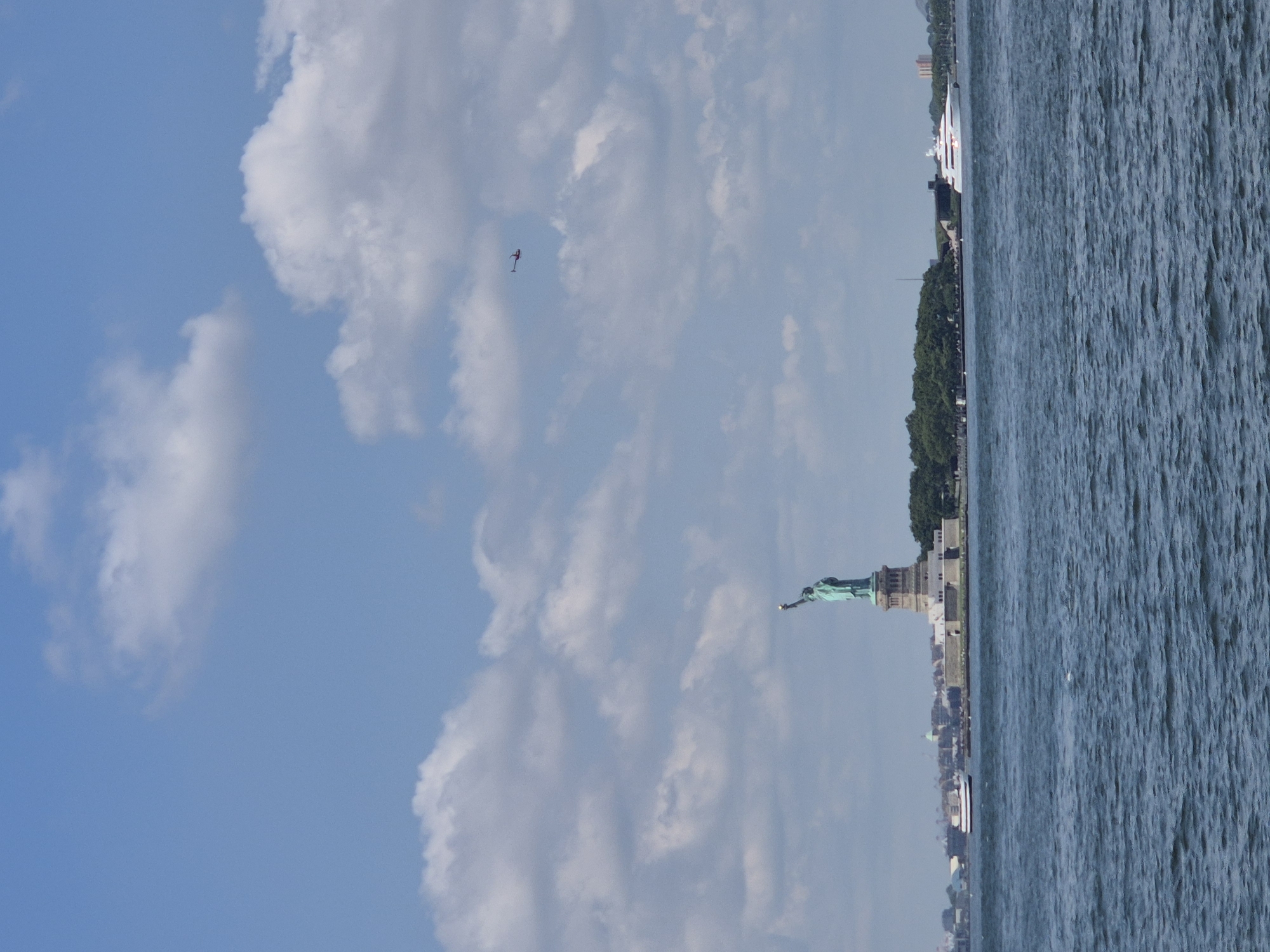 The image captures the iconic Statue of Liberty in New York City, USA, seen from a distance across the water. The statue stands on Liberty Island, with the New York City skyline visible in the background. The sky is a clear blue with scattered white clouds, and the water is a deep blue, with ripples suggesting a gentle breeze. A small red helicopter is visible in the upper right quadrant of the sky, flying in the distance. In the foreground, the water stretches across the bottom of the frame. On the left side of Liberty Island, a ferry can be seen approaching the island. To the right of Liberty Island, a large white yacht is cruising in the distance. The overall scene is a bright, sunny day, offering a classic view of this famous landmark.