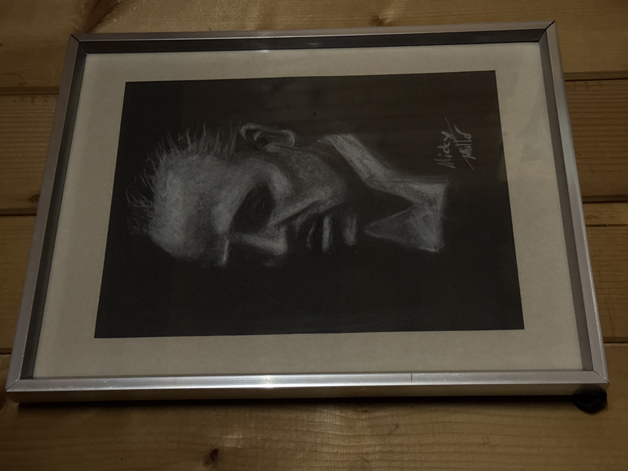 The image shows a framed charcoal drawing of a person's head in profile. The drawing is primarily dark, with white highlights delineating the features of the face. The signature "Micky Mills" is visible in the upper right corner of the drawing. The frame is silver and placed against a wooden paneled wall. Location: Unknown, Unknown.