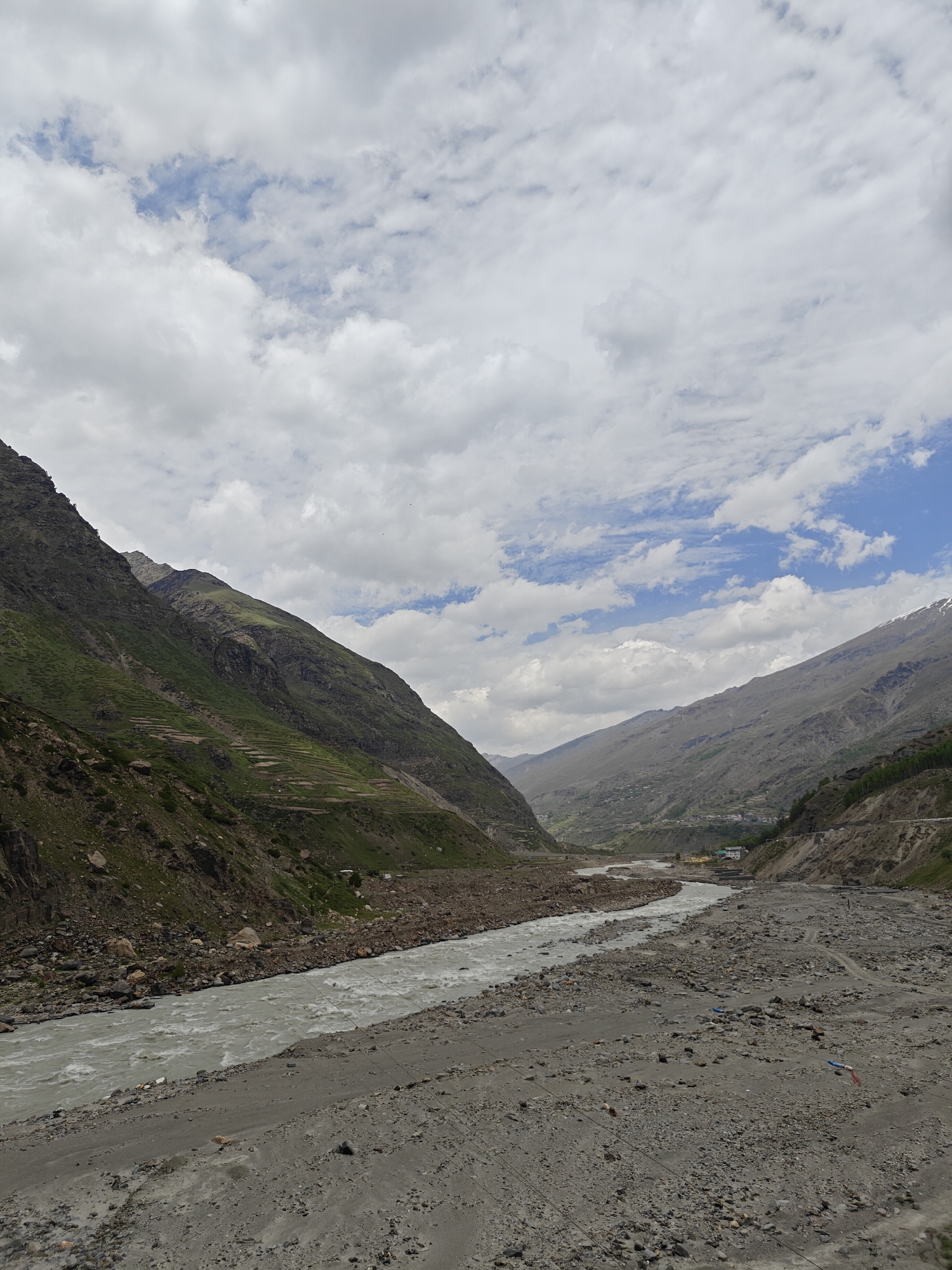 The image depicts a wide, glacial river flowing through a valley in Sissu, India. The river is a murky, light brown color and appears to be flowing swiftly, with whitecaps indicating turbulence. The riverbanks are a mix of rocky terrain and finer sediment, with some evidence of erosion. On the left side of the frame, a steep, green mountain slopes upwards, terraced in sections, suggesting agricultural use. The right side of the valley is also mountainous, with barren, rocky slopes transitioning to greener vegetation at lower elevations. In the distance, the valley narrows, and hints of a settlement with some buildings are visible. The sky above is partially cloudy, with large, fluffy white clouds interspersed with patches of blue sky. The overall impression is of a rugged, natural landscape shaped by the powerful force of the river and the mountainous terrain. No people or specific actions are discernible in the image.