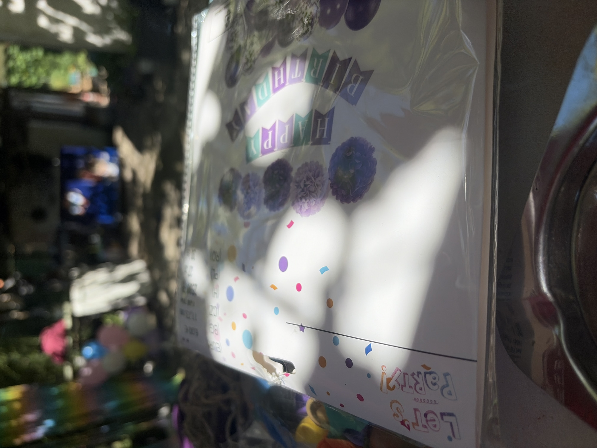 This is an outdoor, close-up, eye-level shot of a packaged party decoration, likely in Vlasatitsa, Bulgaria. The decoration features a "Happy Birthday" banner and "Let's Party!" text, with colorful confetti accents. The packaging is clear plastic, reflecting sunlight and creating shadows on the product. In the blurred background, there are hints of trees, outdoor furniture, and possibly a building structure. The scene is illuminated by natural sunlight.