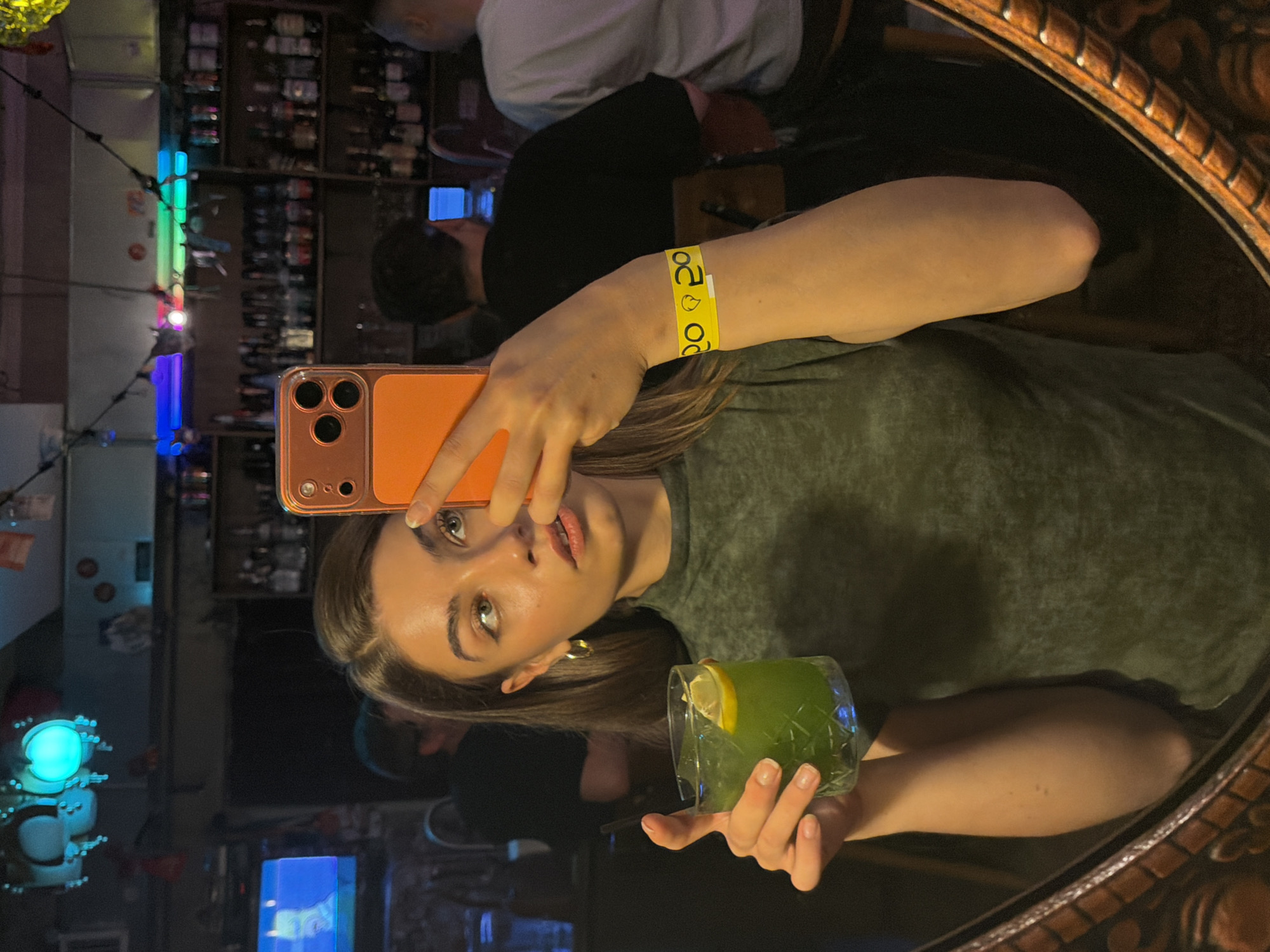 A young woman is depicted in a mirror selfie, holding an orange-cased smartphone in her right hand to capture the image. She has light brown hair and is wearing a dark green top. In her left hand, she holds a clear glass containing a green cocktail with ice and a lemon slice. A yellow wristband with black "20" and face/emoji symbols is visible on her left wrist. Her expression is neutral as she looks towards the phone's camera.
The setting is an indoor space, likely a bar or club in Moscow, Russia, suggested by the blurred background showing shelves filled with bottles. The ambiance is dark, illuminated by various colorful artificial lights including blue, green, purple, and red. The mirror itself has an ornate, carved wooden frame. Other indistinct figures of people can be seen reflected in the background, hinting at a social gathering or event. The time of day appears to be evening or night due to the artificial lighting.