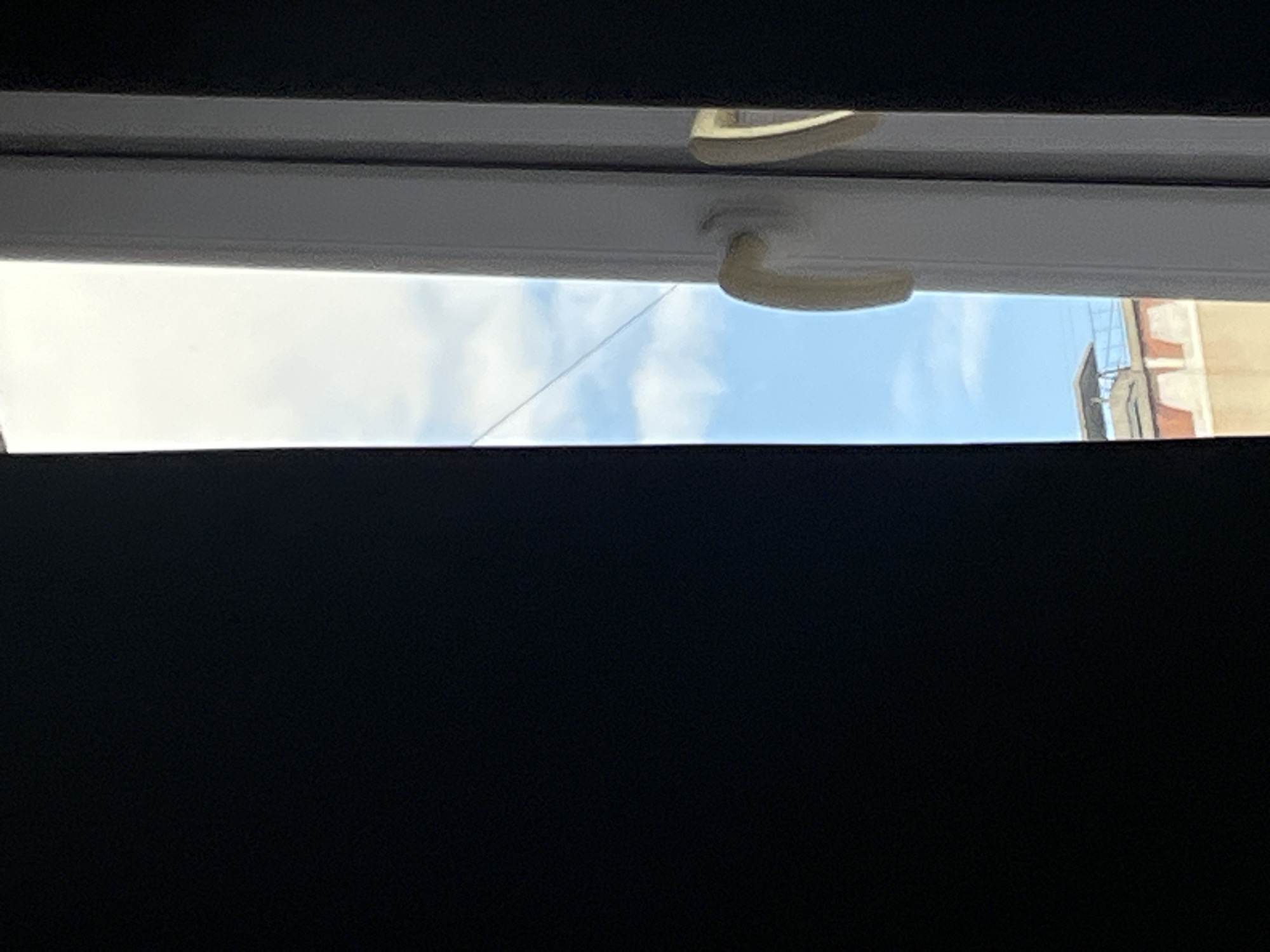 The image shows a view through a white-framed window, with a dark curtain on the left side partially obscuring the scene. Outside the window, the sky is visible with scattered white clouds. The window frame has a cream-colored handle. In the distance, the upper portion of a building with red and beige colored walls is visible, along with a flat roof structure. The image does not contain any information to suggest it is located in Chelyabinsk, Russia.