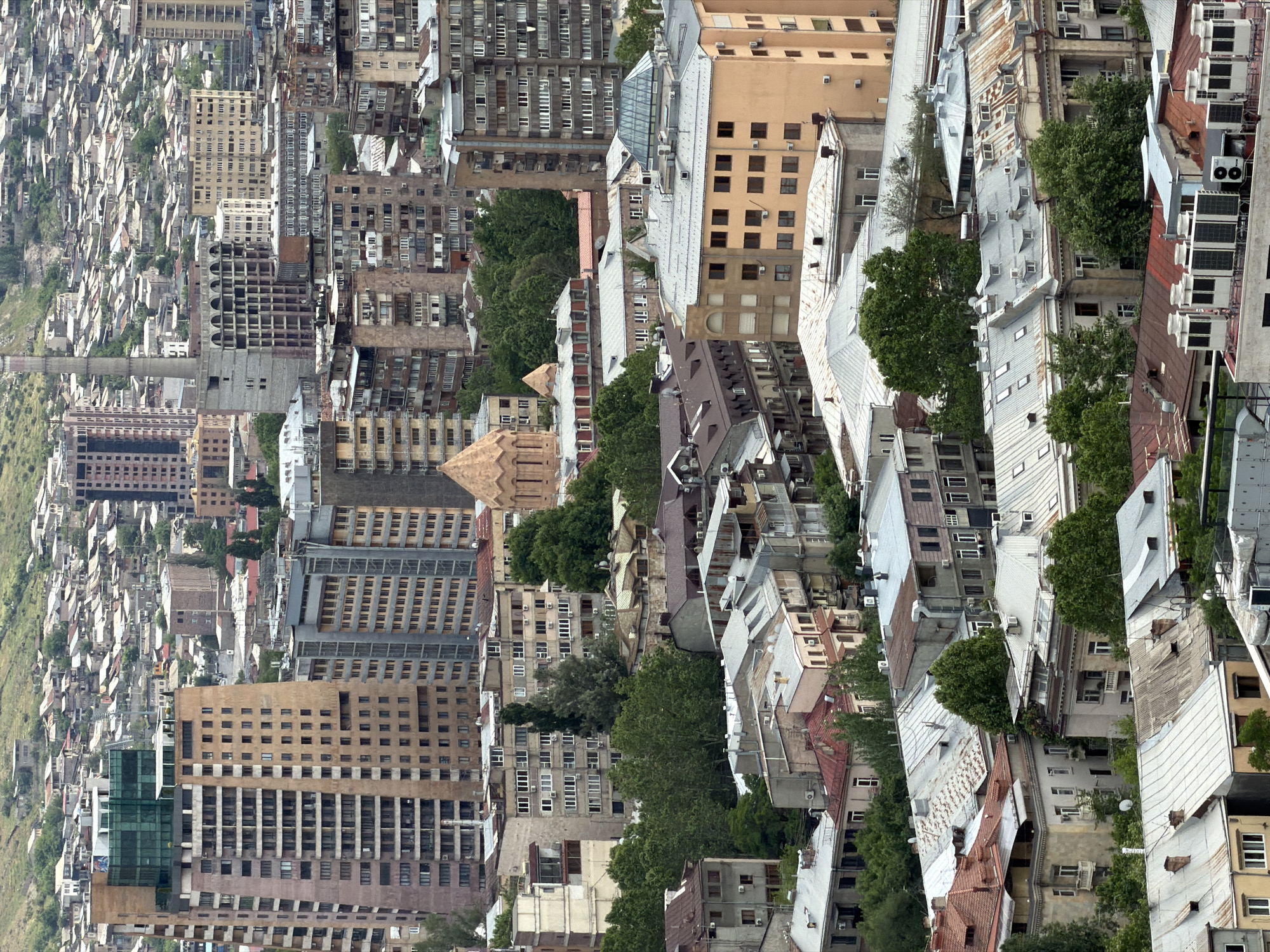 The image presents an aerial view of Yerevan, Armenia. Various buildings, ranging from multi-story apartment complexes to smaller, older structures, dominate the scene. Rooftops vary in color, from shades of red to gray, and many buildings are adorned with air conditioning units. Patches of greenery, including trees, are interspersed throughout the urban landscape. In the distance, the urban landscape fades into what appears to be a more natural, hilly area. The image is rotated counterclockwise.