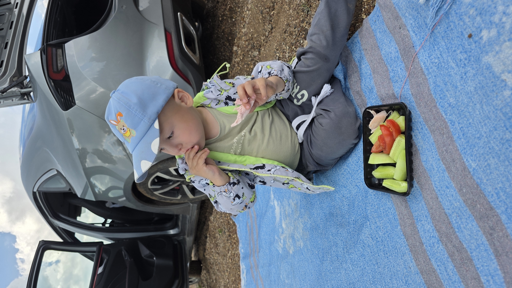 A young child wearing a blue cap with a rabbit design and a grey jacket with a dinosaur pattern is sitting on a blue and grey striped blanket. The child is holding a piece of food, possibly meat, in their right hand and has their left hand near their mouth, suggesting they are eating. In front of the child is a black plastic container with sliced cucumbers and tomatoes. A grey car with its doors and trunk open is parked behind the child. The ground is covered with gravel and dirt. The sky above is cloudy. This scene is taking place outdoors, potentially in a rural or semi-rural area given the presence of gravel and the car.