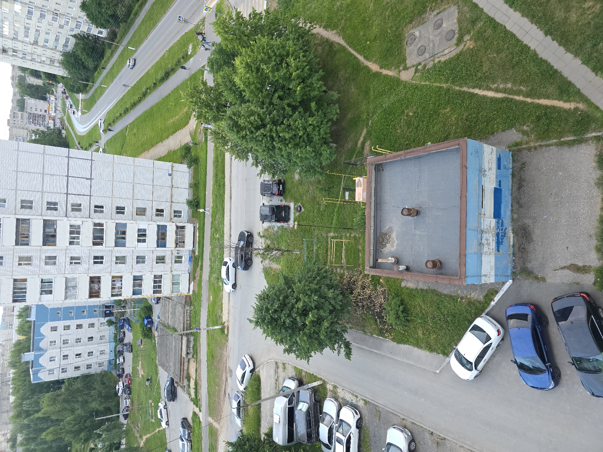 This aerial view captures a residential area in Novocheboksarsk, Russia. In the foreground, a small, utilitarian building with blue and white painted walls and a flat, gray roof stands beside a paved area with parked cars. The cars visible include a white sedan, a blue sedan, and a dark-colored sedan. Further back, a cluster of green trees dominates the landscape, with a yellow metal structure visible behind them. A network of paved paths and grassy areas extends beyond the trees. In the middle ground, a street is lined with parked cars, including a silver van and several other sedans. A light-colored apartment building with many windows rises prominently on the right side of the frame, with another similar building partially visible on the left. Beyond these buildings, a winding road with traffic is visible, bordered by green lawns and trees. In the background, more residential buildings and trees stretch towards the horizon under an overcast sky.