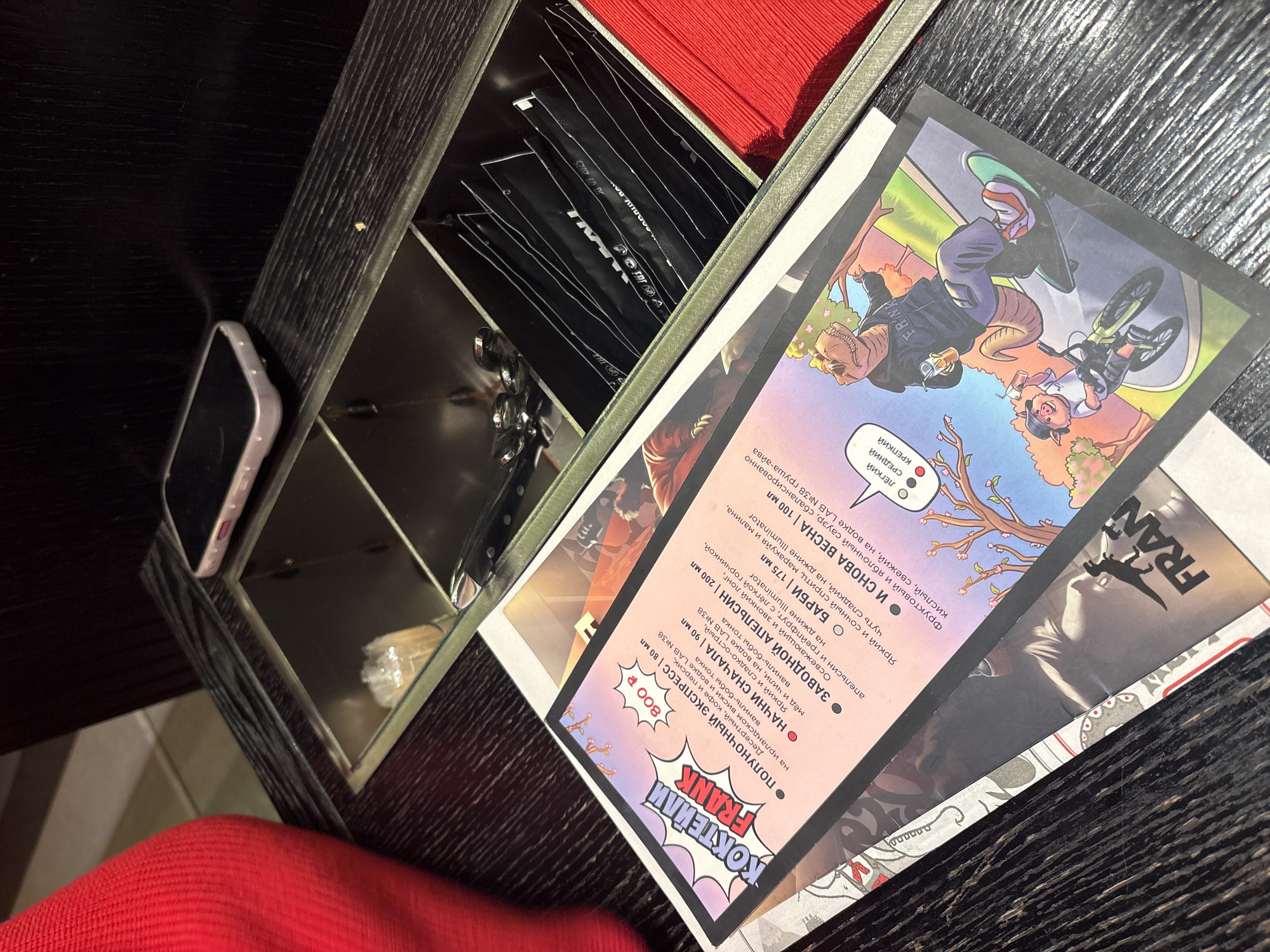 Here is a description of the image:
The image shows a dark, possibly wooden, table with a compartmentalized container on it. The container holds black packaged items (possibly sugar or sweetener), red napkins, and silverware. A menu is placed next to it with an image of characters and a list of cocktails in Russian, including "ПОЛУНОЧНЫЙ ЭКСПРЕСС," "ЗАВОДНОЙ АПЕЛЬСИН," "НАЧНИ СНАЧАЛА," "БАРБИ," and "И СНОВА ВЕСНА," along with prices. A phone lies next to the container. The edge of a red garment is visible in the bottom left corner, suggesting someone is seated at the table. The context suggests a cafe or restaurant setting. The cocktails in Russian suggest the location is likely in Russia, potentially Saint Petersburg based on the additional information provided.
