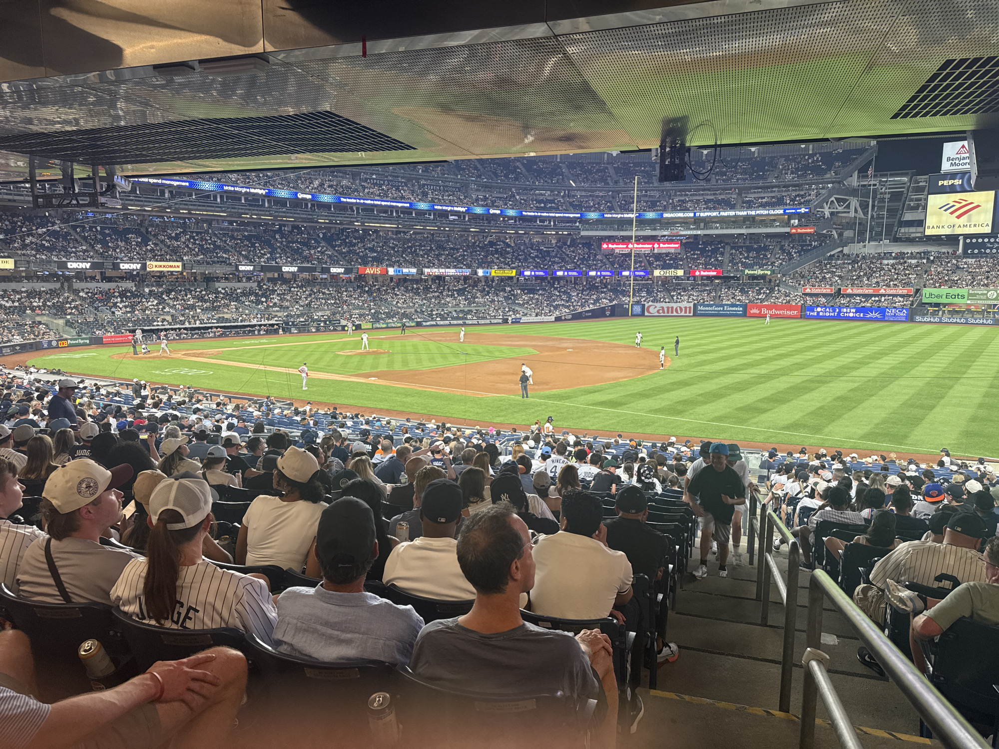 This image depicts a night baseball game in progress at Yankee Stadium, located in The Bronx, New York City, United States of America. The scene is viewed from an elevated seating area, looking down towards the playing field and across to the upper decks of the stadium.
On the brightly illuminated baseball field, a game is actively being played. A batter is visible at home plate, with a catcher crouched behind and an umpire standing nearby. A pitcher is positioned on the pitcher's mound, while several fielders are dispersed across the dirt infield and the striped green outfield grass. Additional players or coaches are present in the dugout areas. A tall foul pole is visible in the right field.
In the foreground, numerous spectators occupy dark stadium seats, all facing the field. Many attendees are wearing baseball-themed apparel, including New York Yankees caps and jerseys, with one person's jersey displaying the name "Judge." A person in the lower left holds a beverage can. Towards the right, an individual is observed walking up a concrete stairway between rows of seats, with metal railings alongside.
The stadium's upper tiers are also populated with spectators. Above the distant stands, multiple digital display boards and ribbon screens showcase various advertisements and sponsor logos. Visible brands include "Bank of America," "Pepsi," "Uber Eats," "Benajmin Moore," "DKNY," "Kikkoman," "Avis," "Budweiser," "State Farm," "Canon," "Montefiore Einstein," "Poland Spring," "Aspen Vodka," and "Oris." The underside of the overhead structure features a perforated metal ceiling.