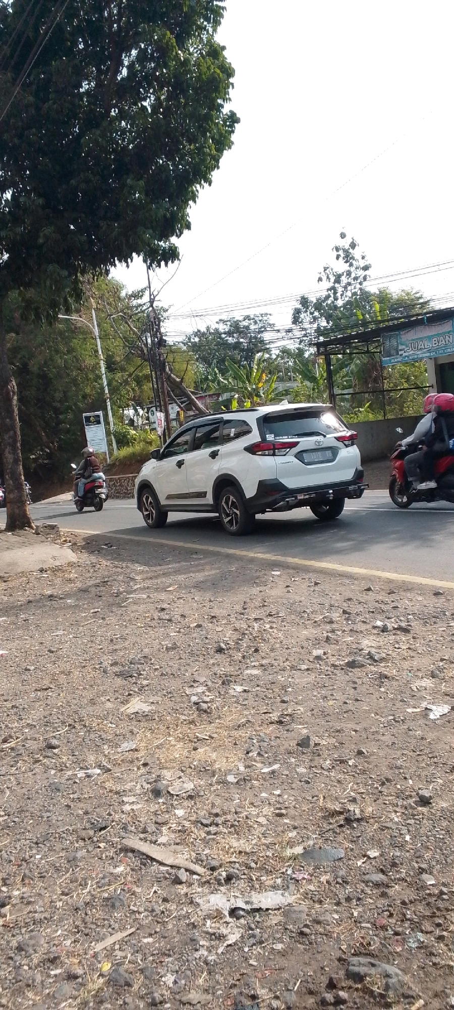 The image depicts a street scene in Cilaku, Indonesia, on a bright, clear day. A white SUV is driving on the road, positioned in the center of the frame. To the left of the SUV, a person is riding a red scooter. Further to the right, another person is riding a red scooter, also on the road. The road itself is asphalt with a yellow line marking the lane. The foreground is a gravel and dirt shoulder littered with debris. In the background, there are trees, utility poles with numerous wires, and a building with signage. One sign, partially visible, reads "JUAL BAN" (which translates to "TIRE SALE" in Indonesian). The sky is bright and overcast, suggesting a sunny day.