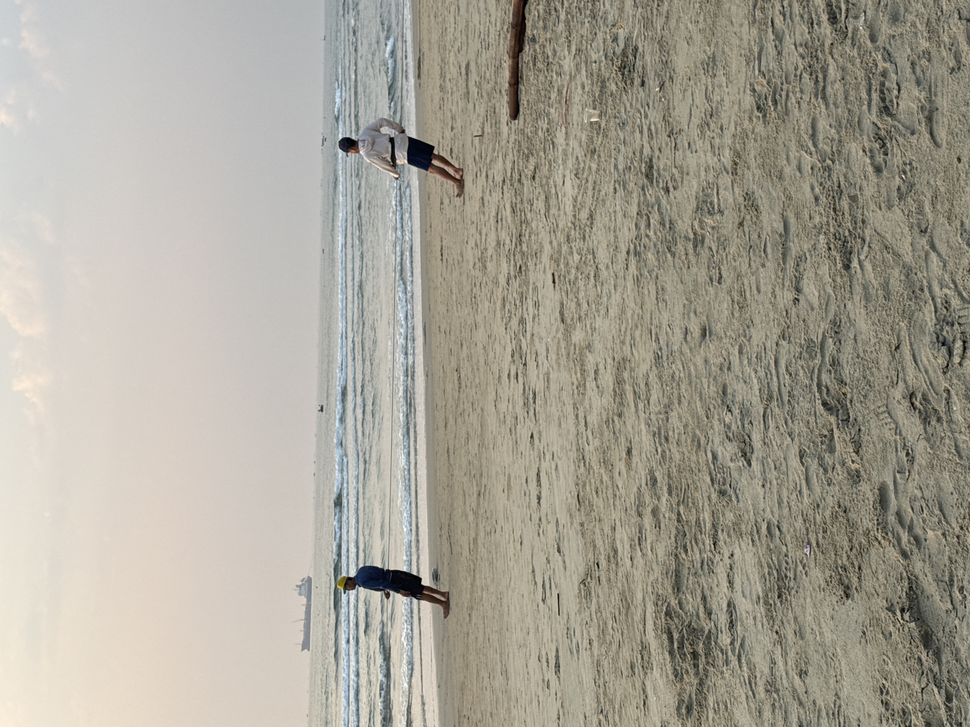 The image depicts a beach scene in Đà Nẵng, Vietnam. Two people are visible on the beach, one wearing a white shirt and dark shorts, and the other wearing a dark shirt, dark shorts, and a yellow hat. The ocean is visible in the background, with small waves approaching the shore. A large ship is visible in the distance. The sand is light-colored and textured, with some debris scattered on the surface. A wooden post is also visible on the sand.