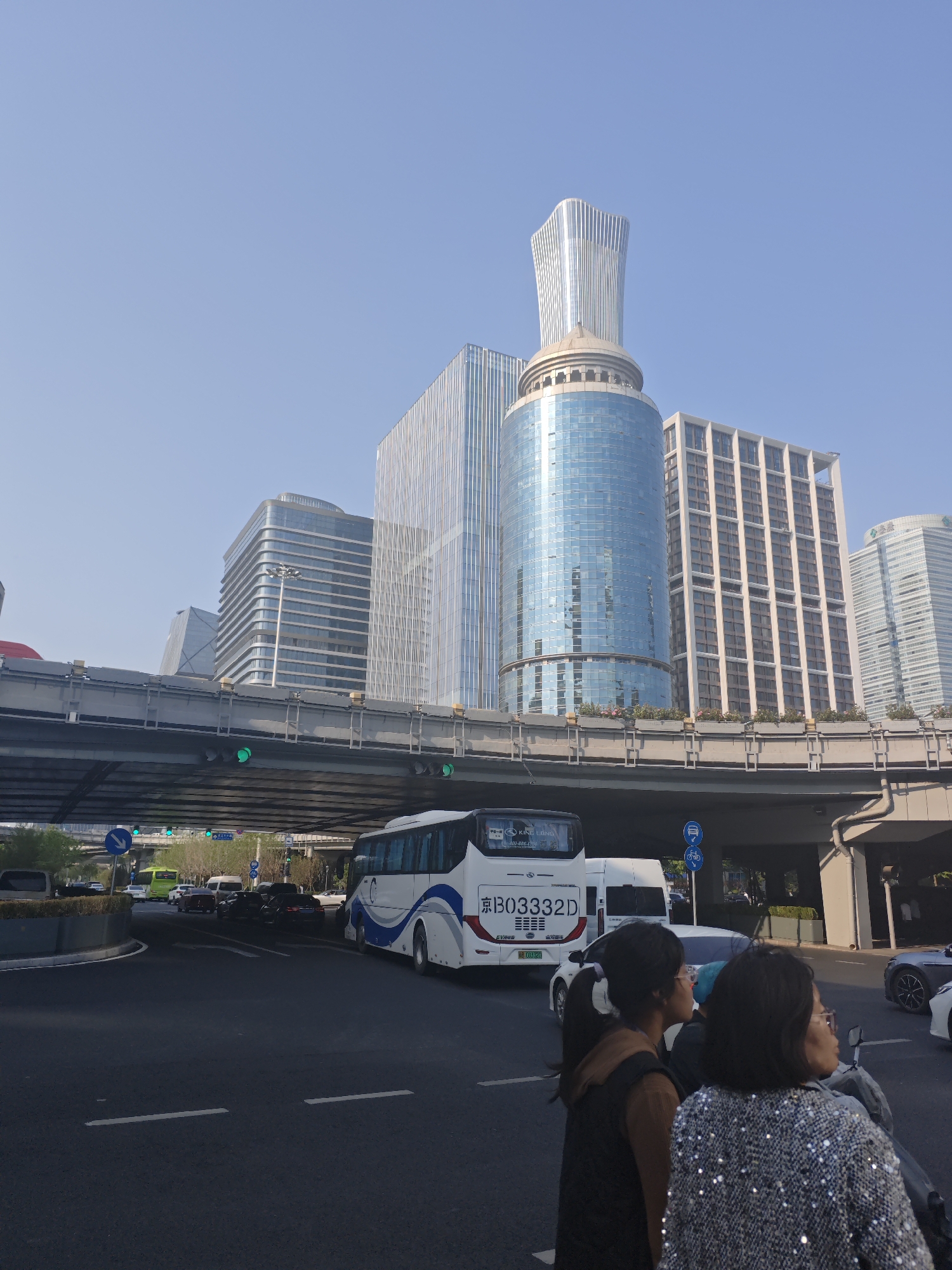 This image presents a vibrant urban street scene in Beijing's Chaoyang District, China, captured during a clear daytime. The view looks up towards a complex architectural landscape and a busy intersection.
Dominating the background are several modern skyscrapers under a bright, cloudless sky. A prominent building features a distinctive fluted, tapering top above a cylindrical, glass-paneled base, reflecting the blue sky. Other surrounding buildings showcase varying architectural styles, from sleek glass facades to structures with rectilinear window patterns in lighter tones.
In the mid-ground, a substantial concrete overpass or elevated road structure spans horizontally across the frame, with metal railings and supporting beams visible. Green traffic lights are visible both beneath and on the elevated road, indicating active traffic flow.
The foreground features a multi-lane road where several vehicles are present. A large white and blue public bus, bearing the license plate "京B03332D" and the brand "KING LONG," is positioned on the right, possibly stopped or moving slowly. Other vehicles include various cars (white, black, grey) and a white van. Road markings, including dashed white lines, are visible on the asphalt. Blue circular road signs, one indicating a left turn and another depicting bicycle and scooter icons, are also present.
Two individuals are captured in the immediate foreground, seen from behind. One person has dark hair tied back, wearing a brown top and a dark vest. Beside them, another person with shorter dark hair wears glasses and a shimmering, sequined top. They appear to be waiting or moving with the traffic, likely on a scooter or similar two-wheeled vehicle. The scene depicts a typical bustling city environment.